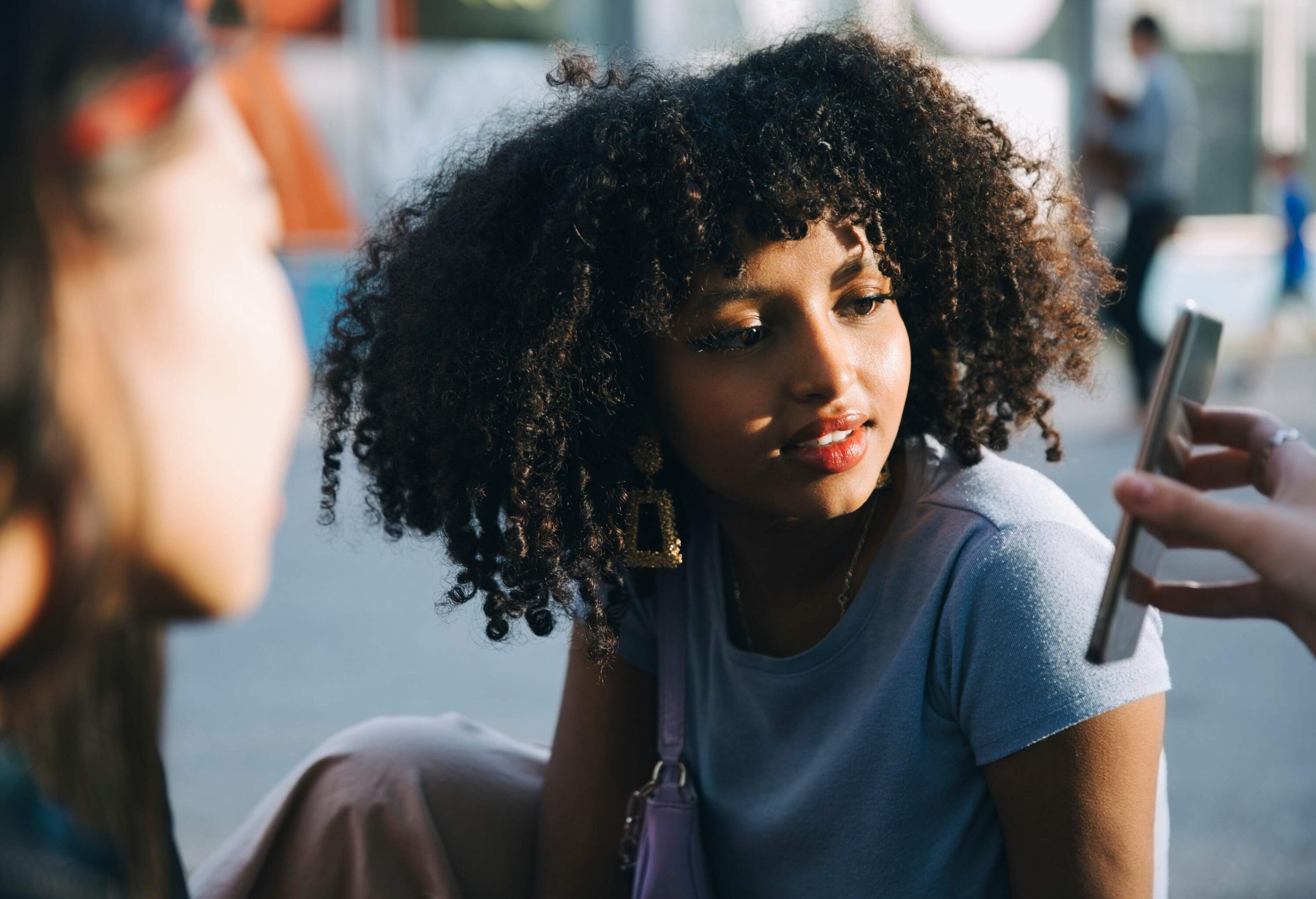 A young woman with kinky hair staring at a smartphone that another person is showing her.