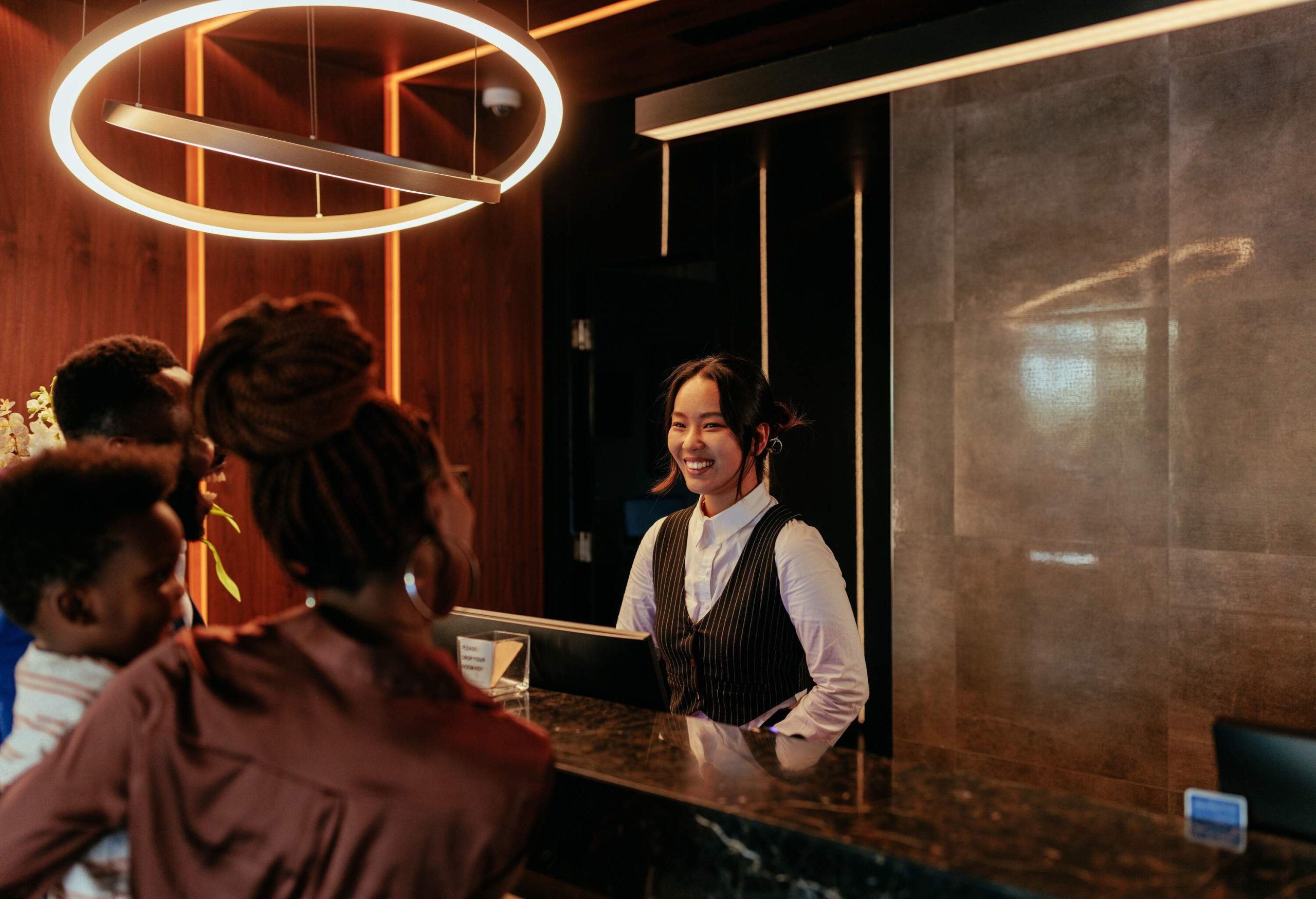 A young Asian receptionist is at the front desk of a luxurious hotel talking to her African American hotel guests.