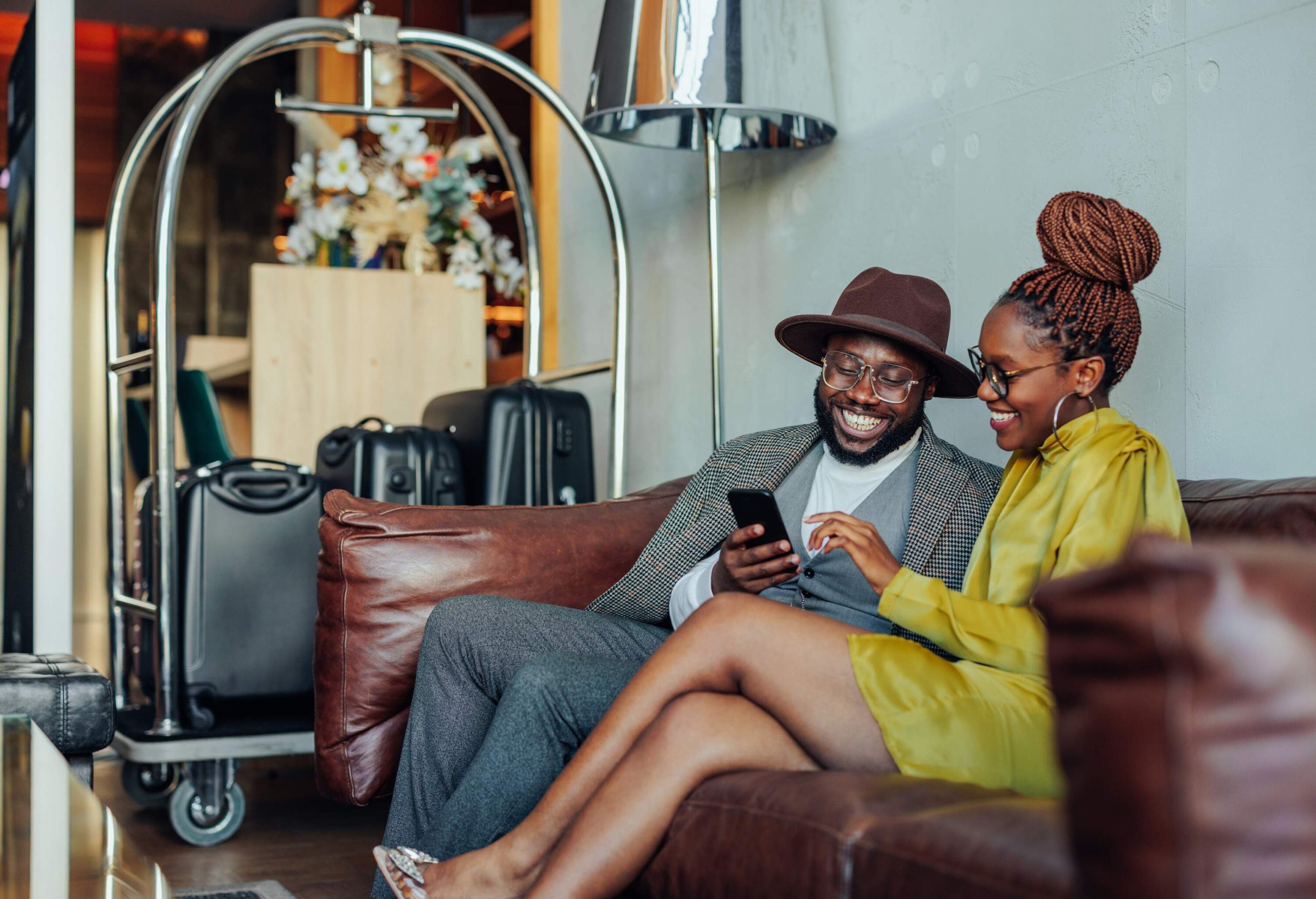A man and a woman sitting with a phone on a couch in a hotel lobby with their suitcases next to them
