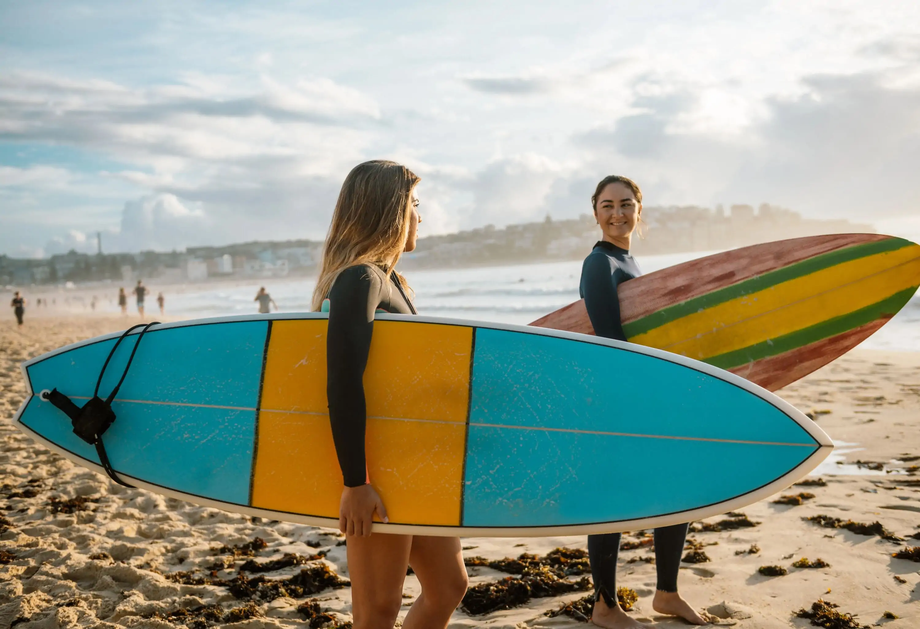 Two women smile at each other as they carry their surfboards on the beach.
