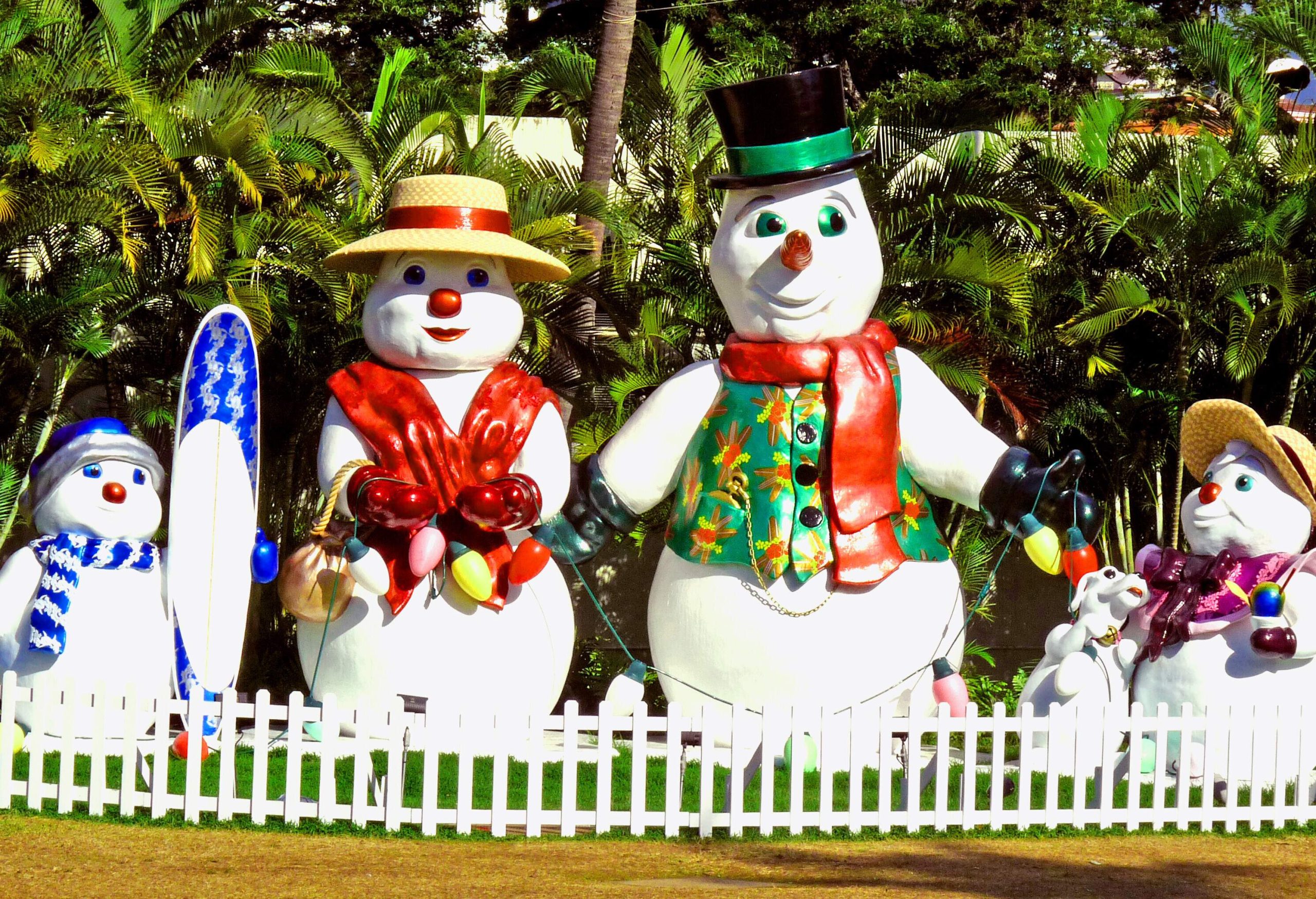 A family of colourful snowman sculptures on a meadow with white fence and palm plants in the back.