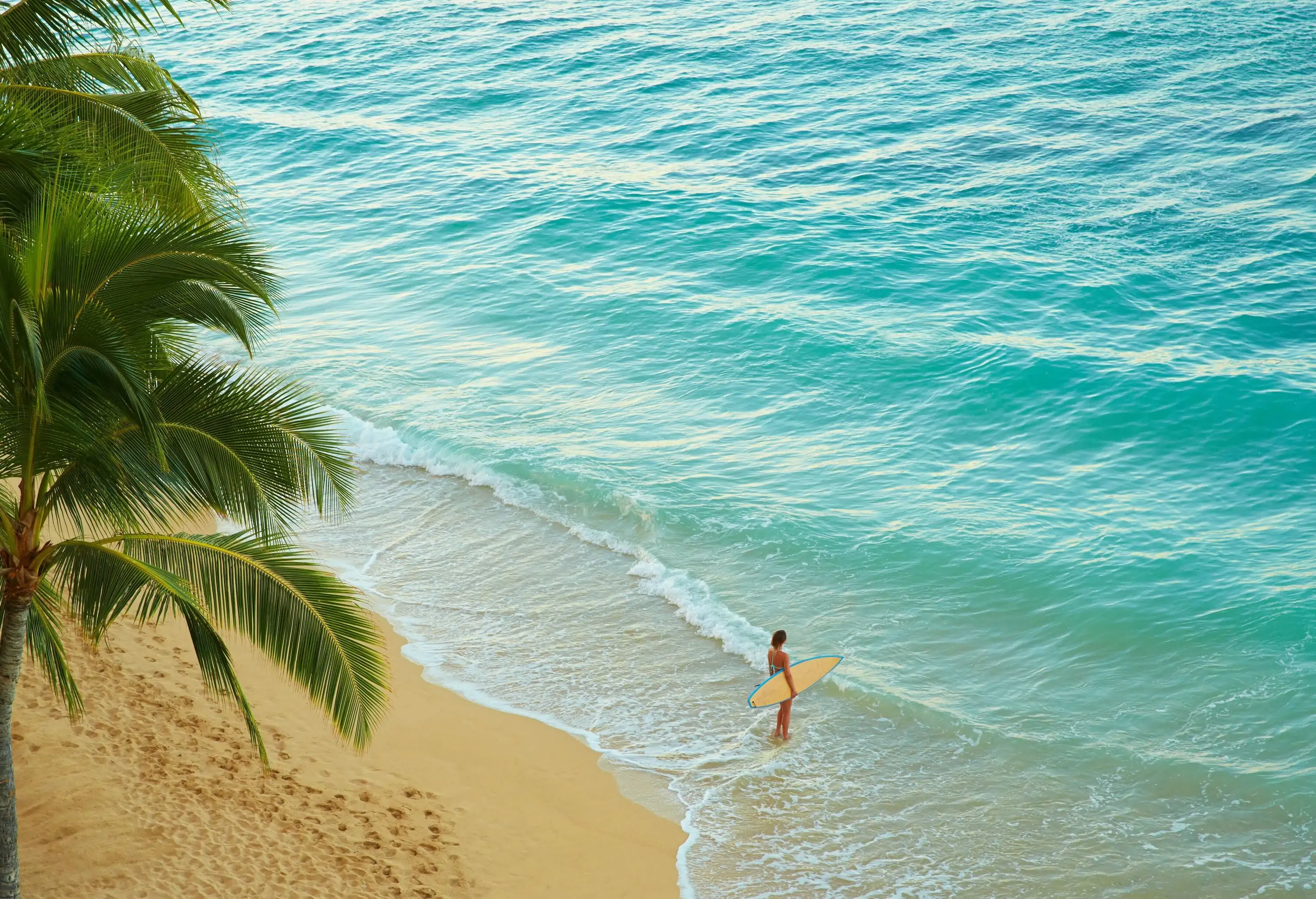 A woman standing on the beach with a surfboard while the waves smash on her feet.