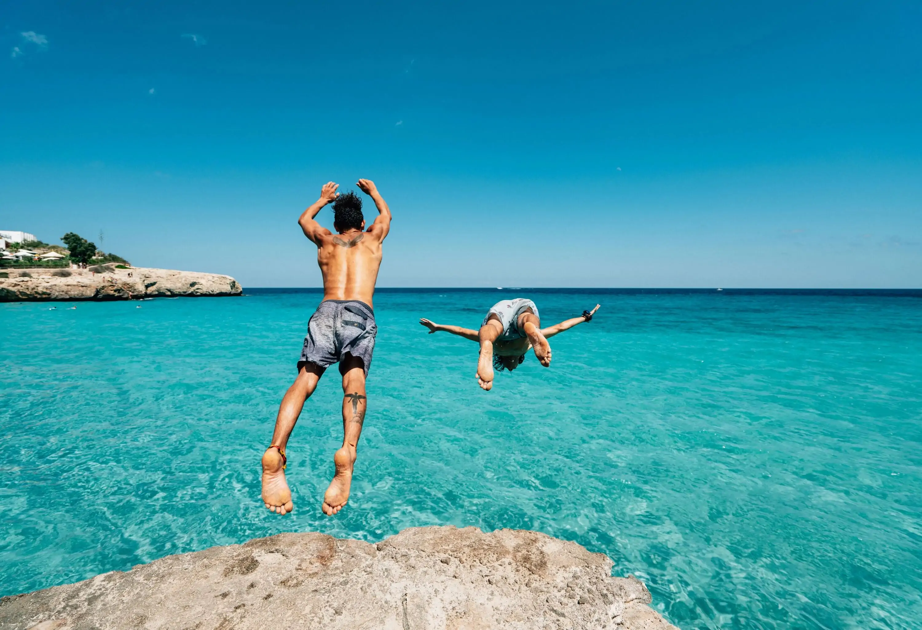 Two men dive into a turquoise ocean from a coastal cliff.