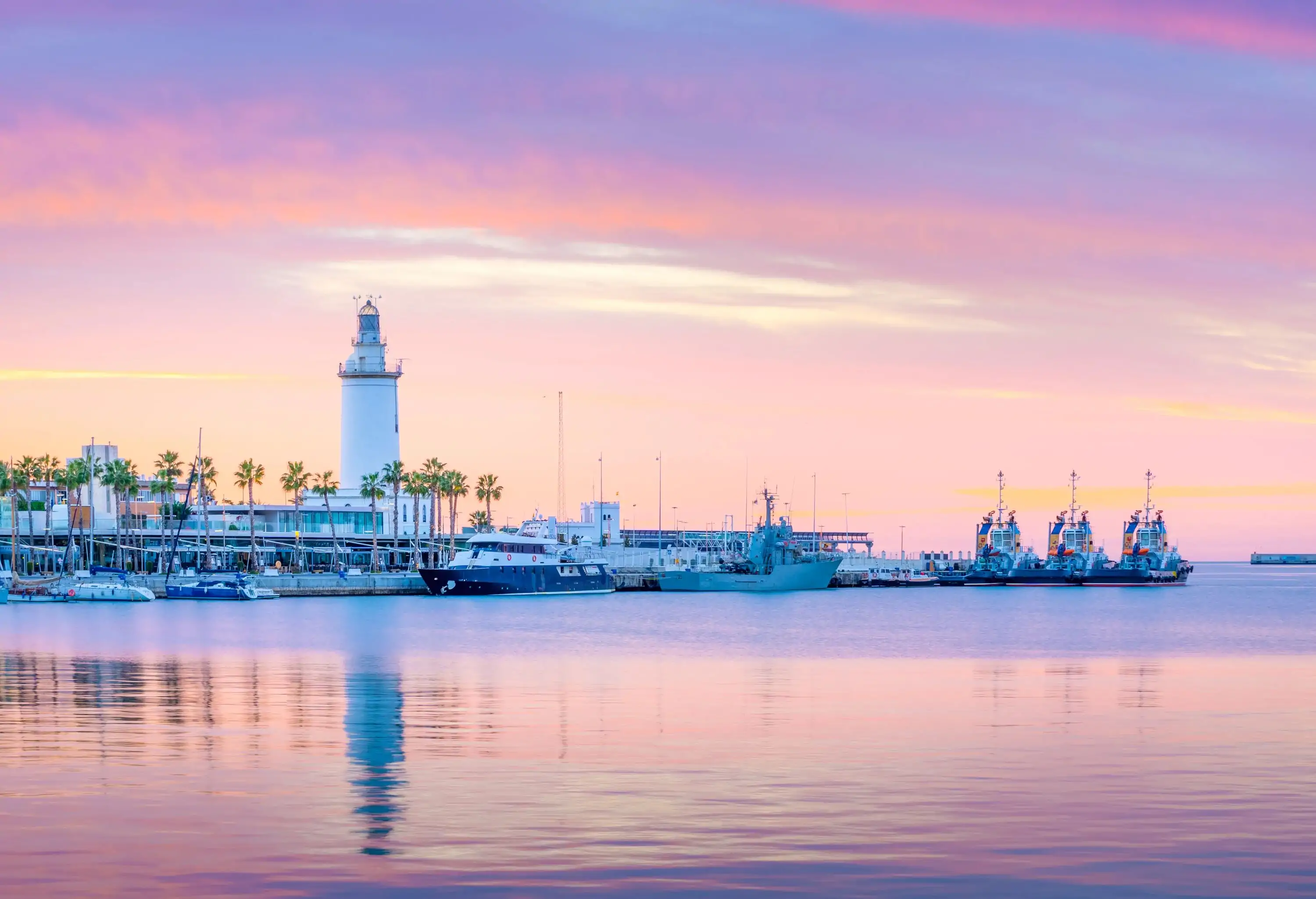 A lighthouse towering above a row of palm trees on the shore and boats in the harbour.