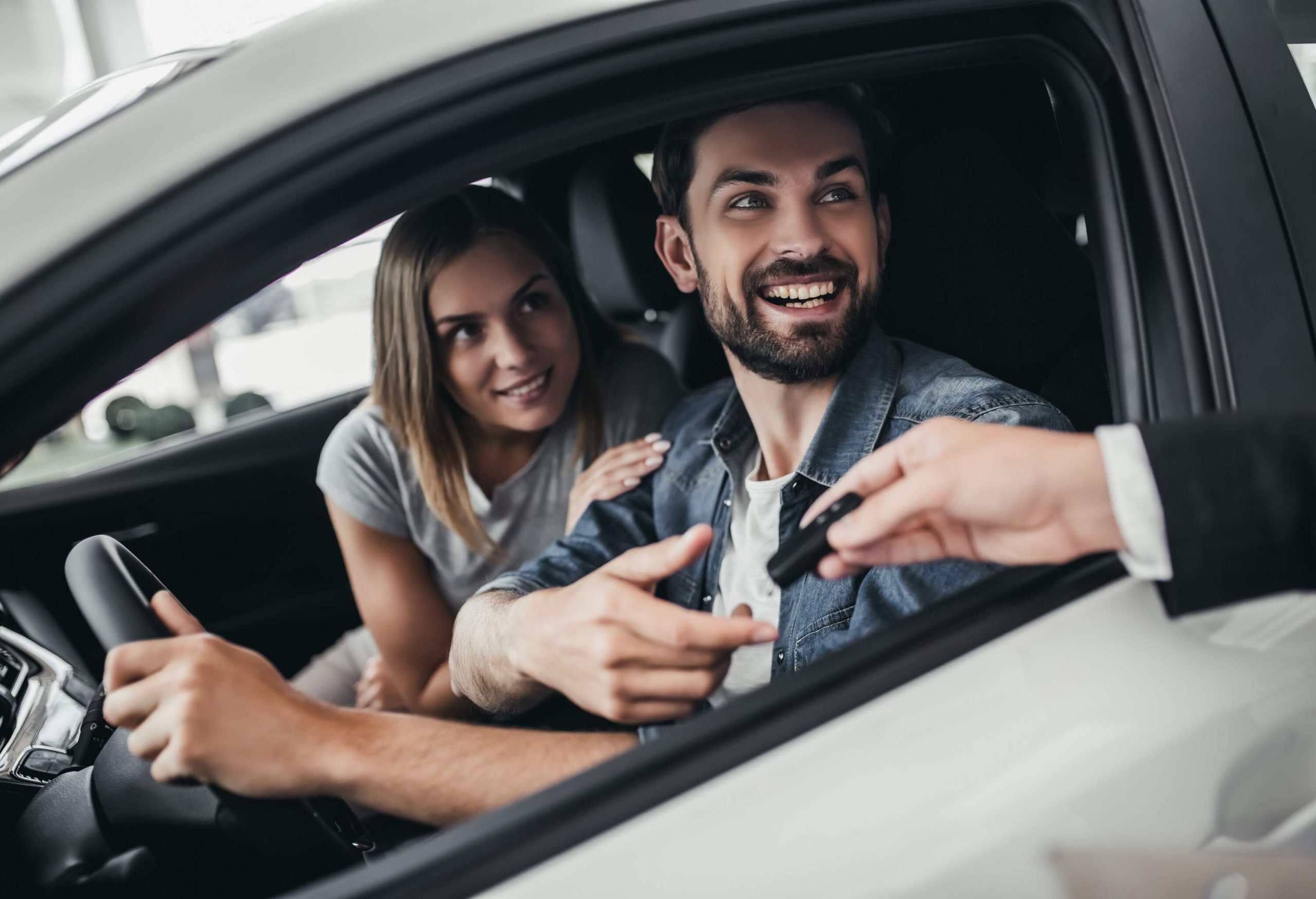 Two happy individuals inside a car look out the driver's window.