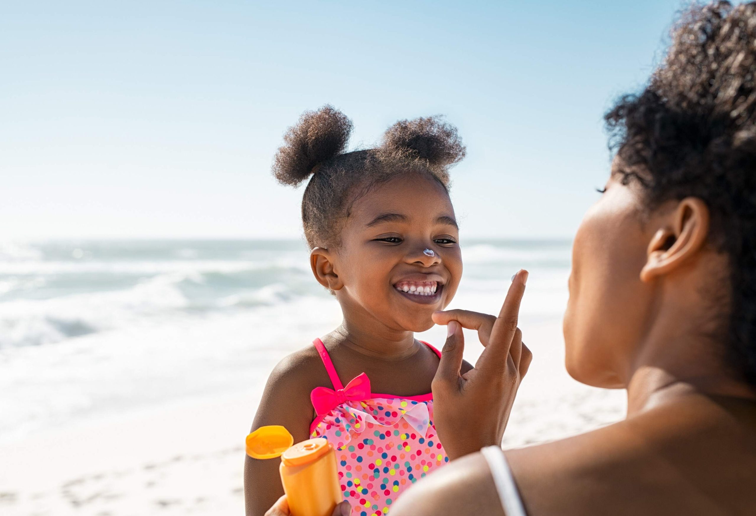 A mother applying a small dab of sunscreen on her daughter's nose in a playful manner.