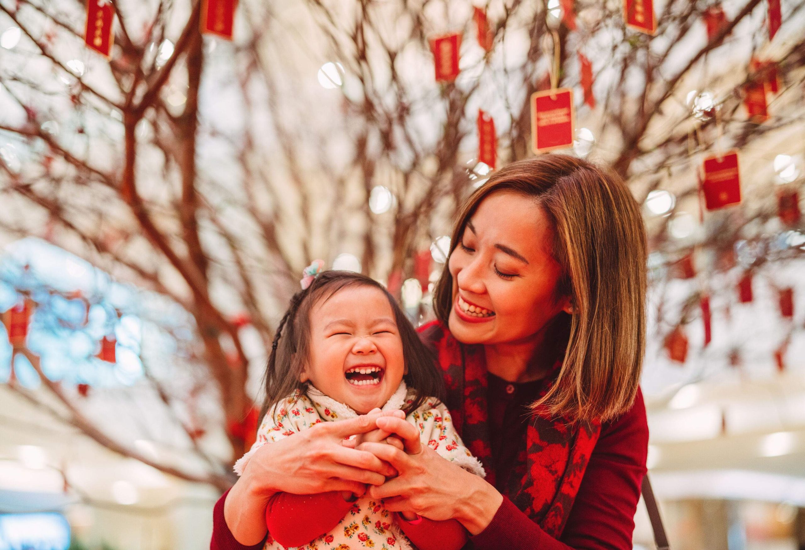 Mother making kung hei fat choy hand gesture with her lovely daughter joyfully in front of the peach blossom tree in Chinese New Year