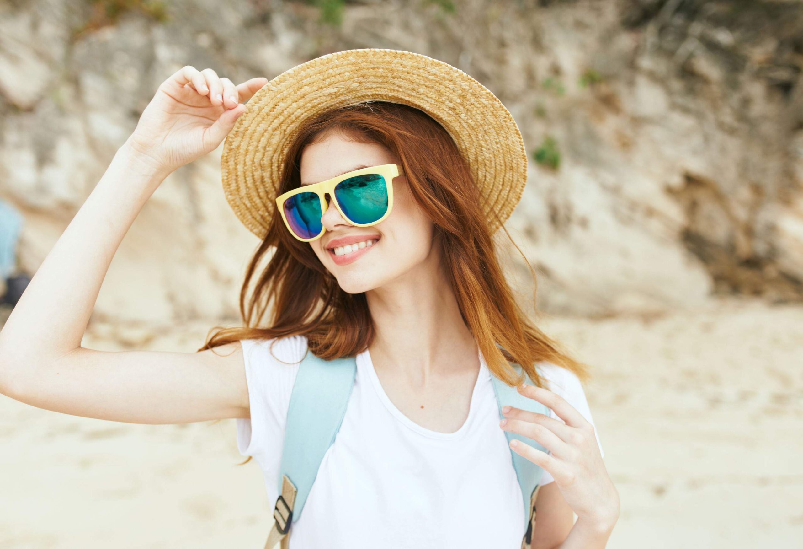 A happy young woman standing on the beach sporting a straw hat, a white shirt, and yellow sunglasses.