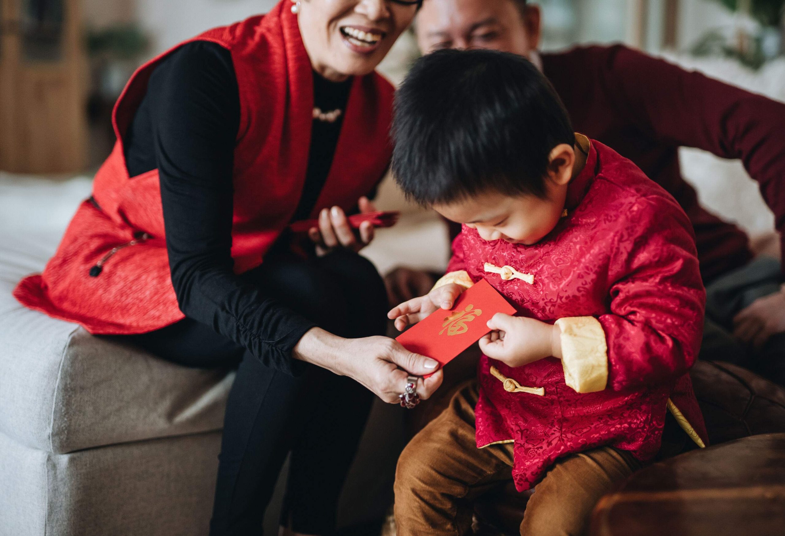 Small child dressed in red traditional Chinese costume receives red envelops (lai see) with both hands from grandparents joyfully in Chinese New Year
