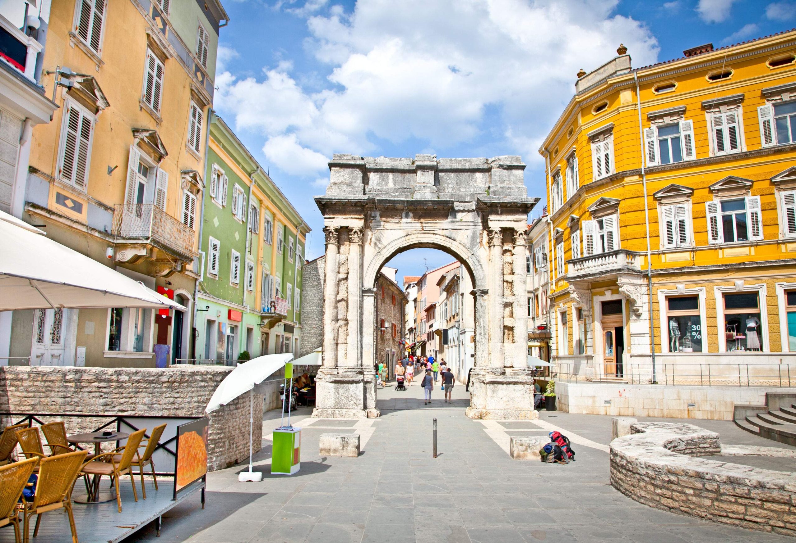 As they make their way along a street lined with bright buildings, individuals pass beneath an ancient archway.