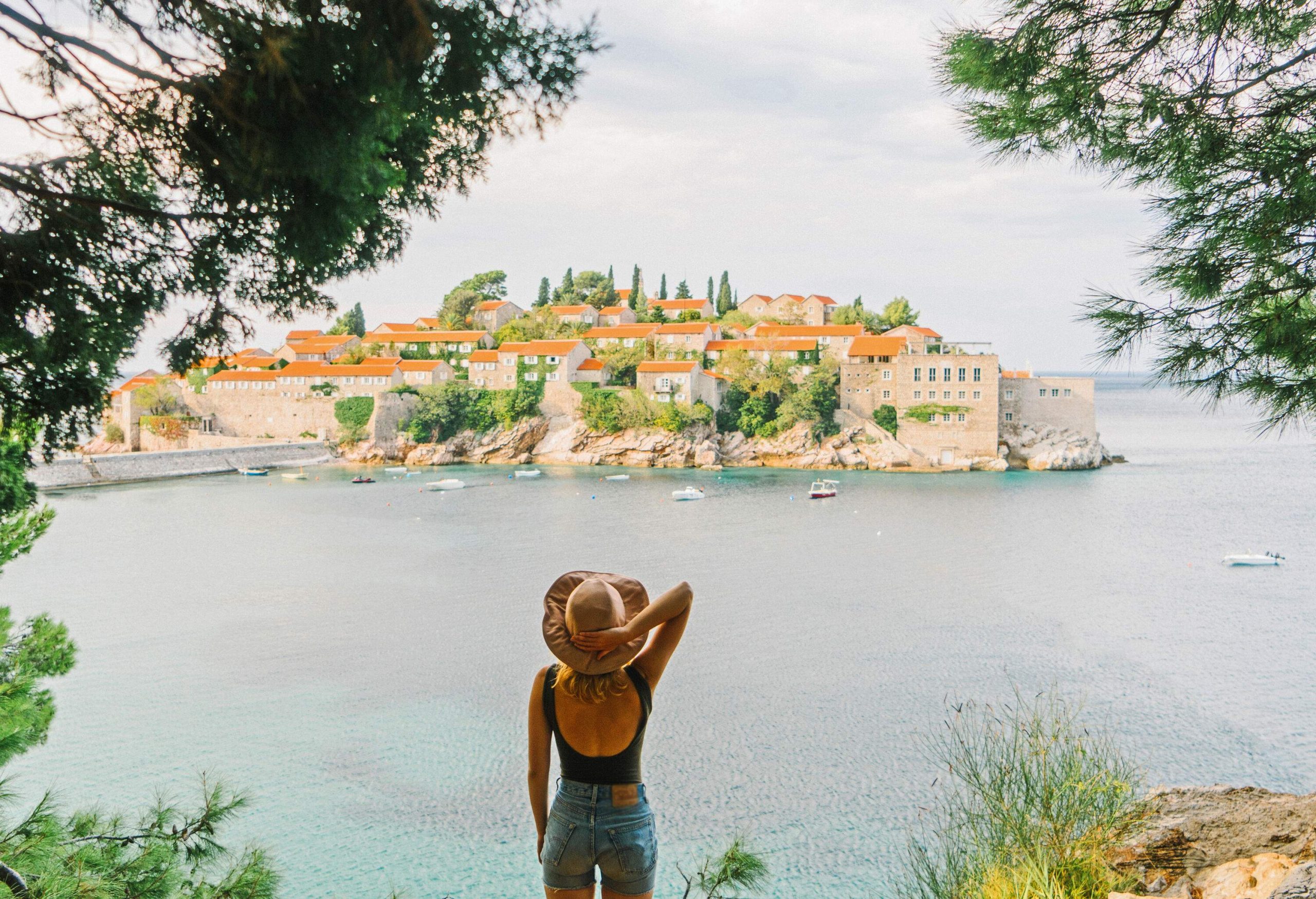 A captivating woman stood poised against the backdrop of the iconic Sveti Stefan island, a picturesque islet adorned with rustic stone buildings and embraced by crystal-clear turquoise waters.