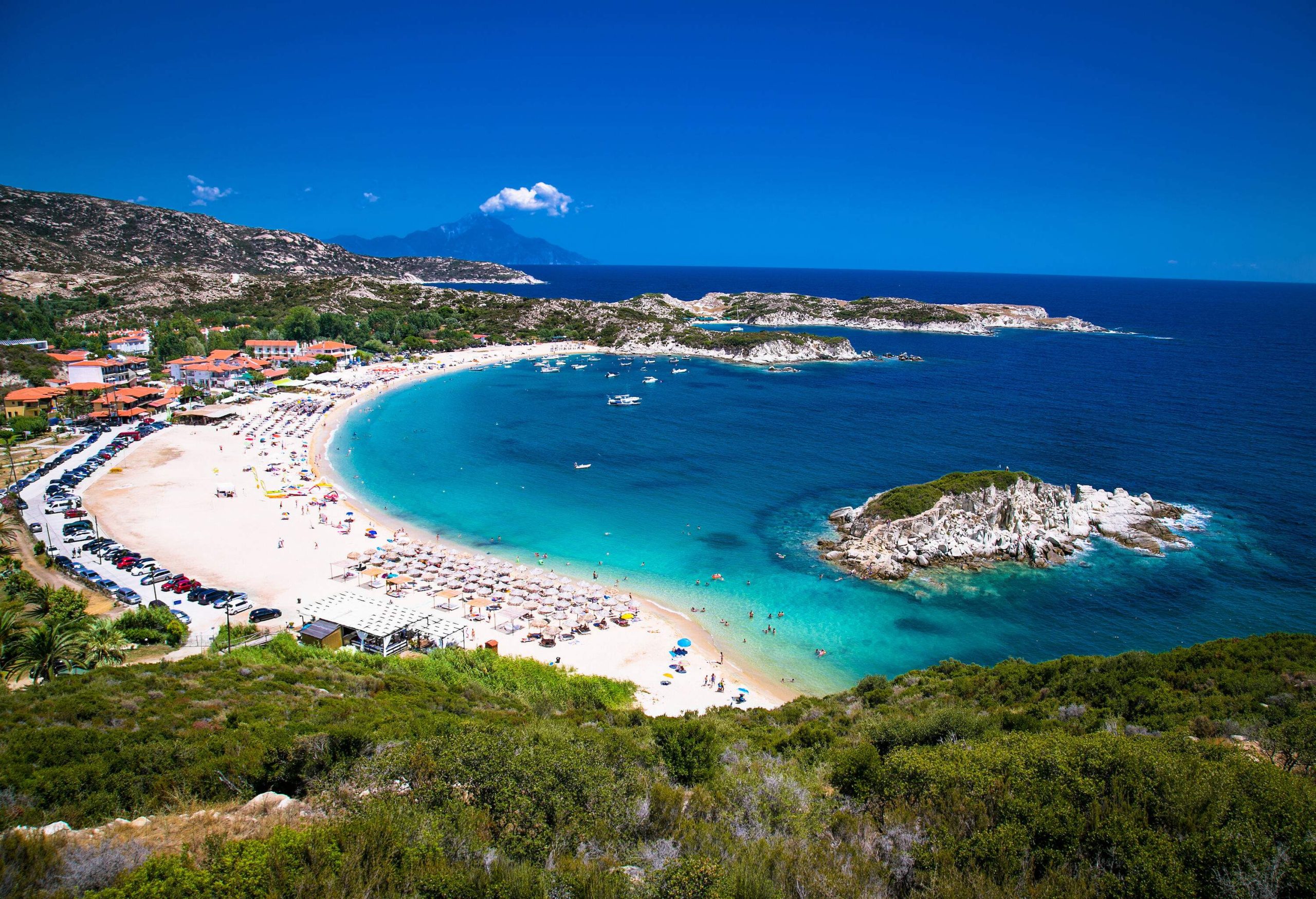 Several rock formations on the seafront and a line of parasols with beach beds enhance the scenic attractiveness of the azure sea, the white sand, and the surrounding vegetation.