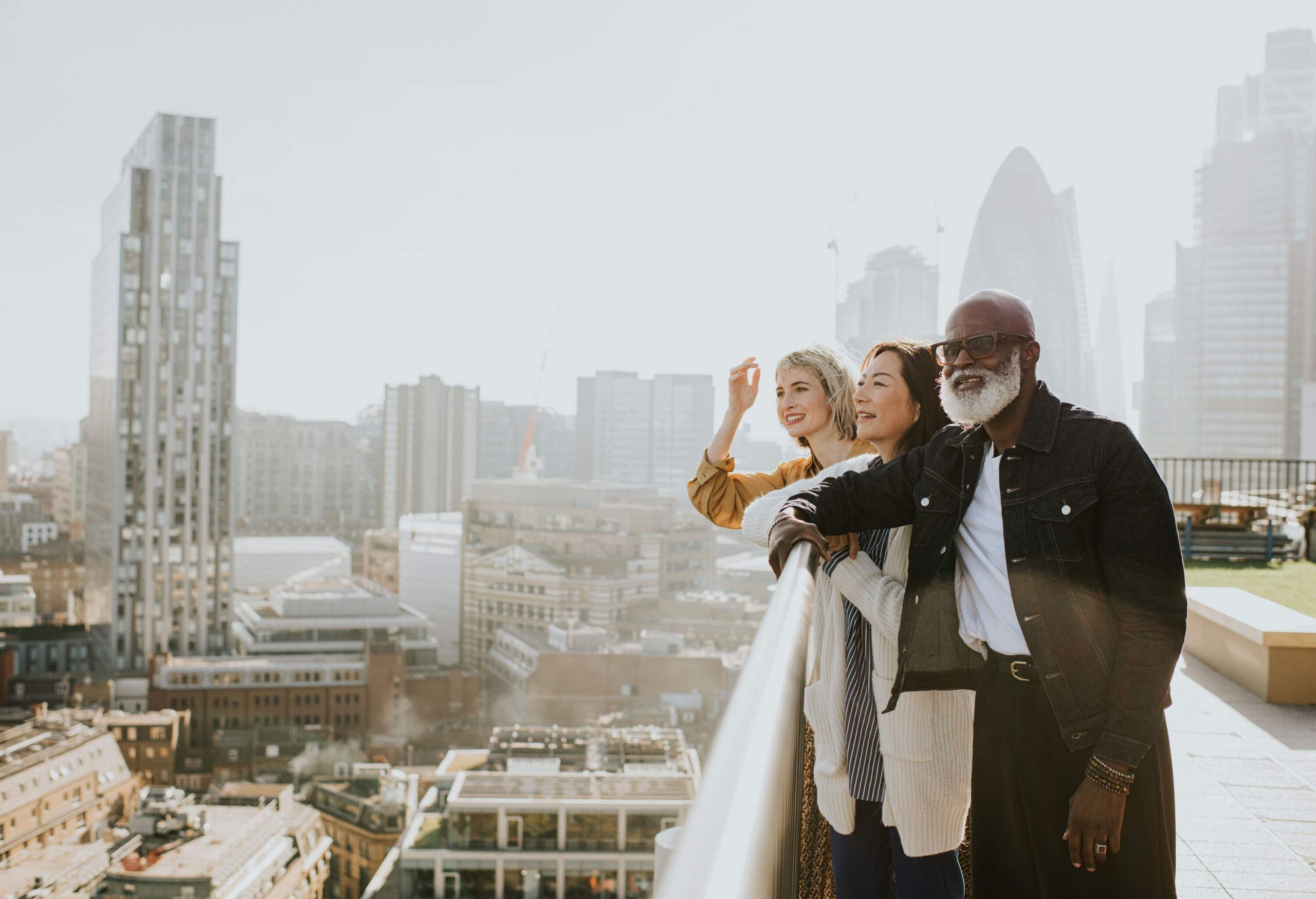 Three people stand on a rooftop / balcony and look out at an epic, sunny cityscape. They admire the view, smiling and pointing and discussing the sights.