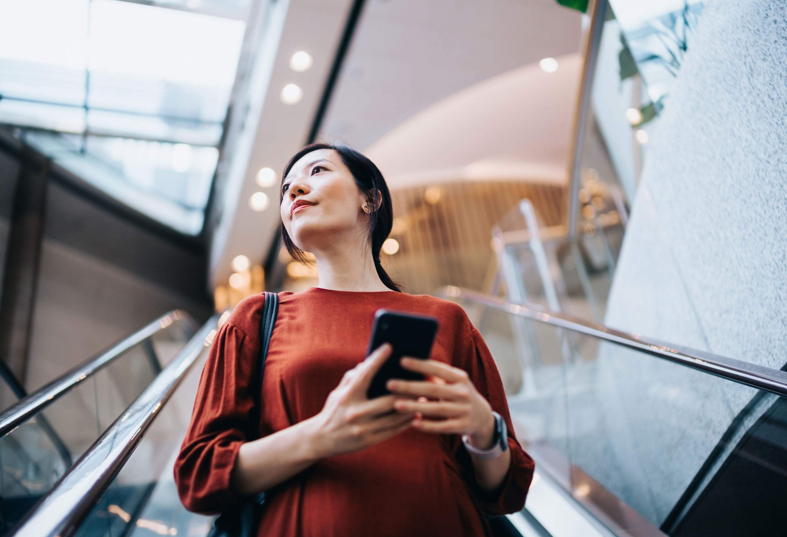 A young Asian woman confidently holds her smartphone with both hands while riding on an escalator, gazing thoughtfully into the distance.