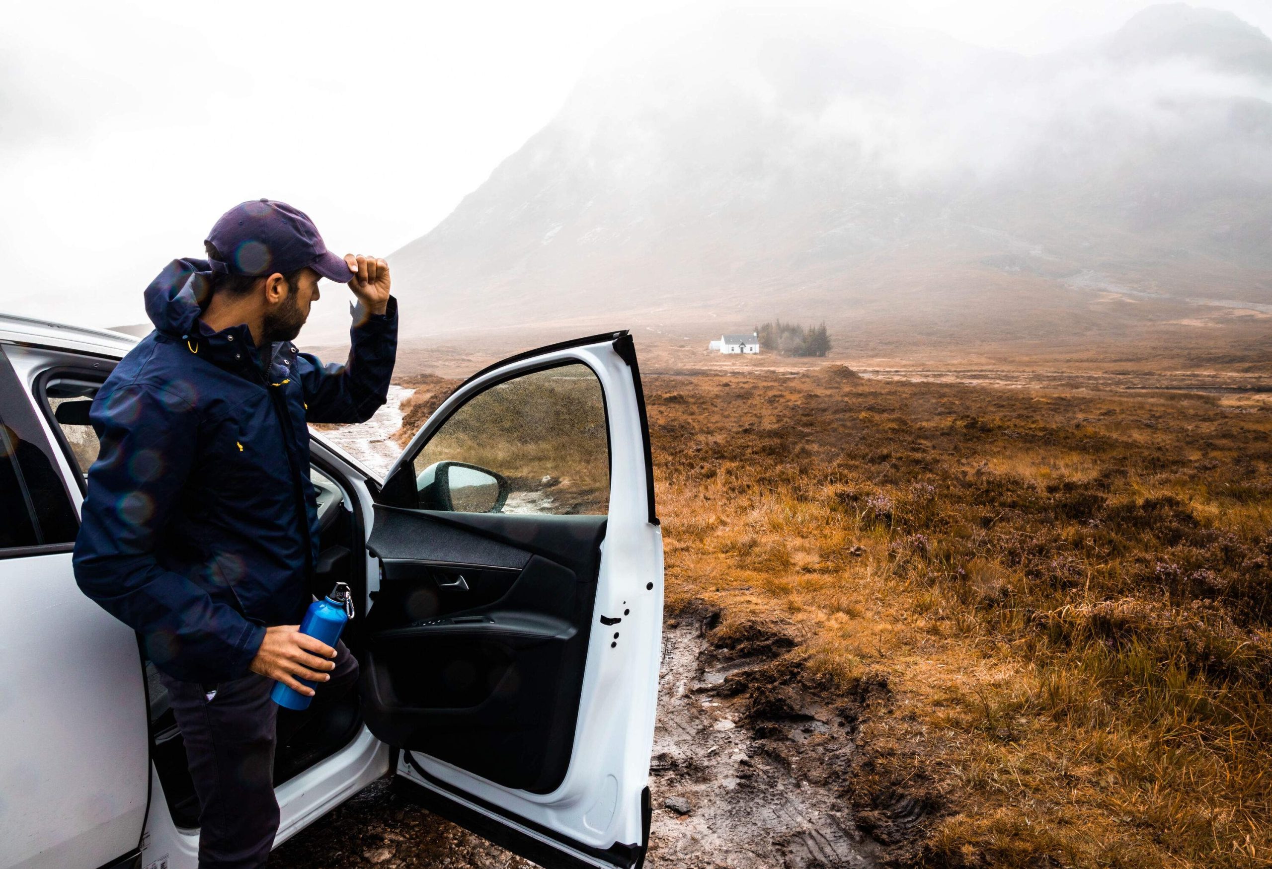 Amidst the challenging conditions of mud and rain, a determined individual clad in a blue windbreaker emerges from a white car, equipped with a flask.