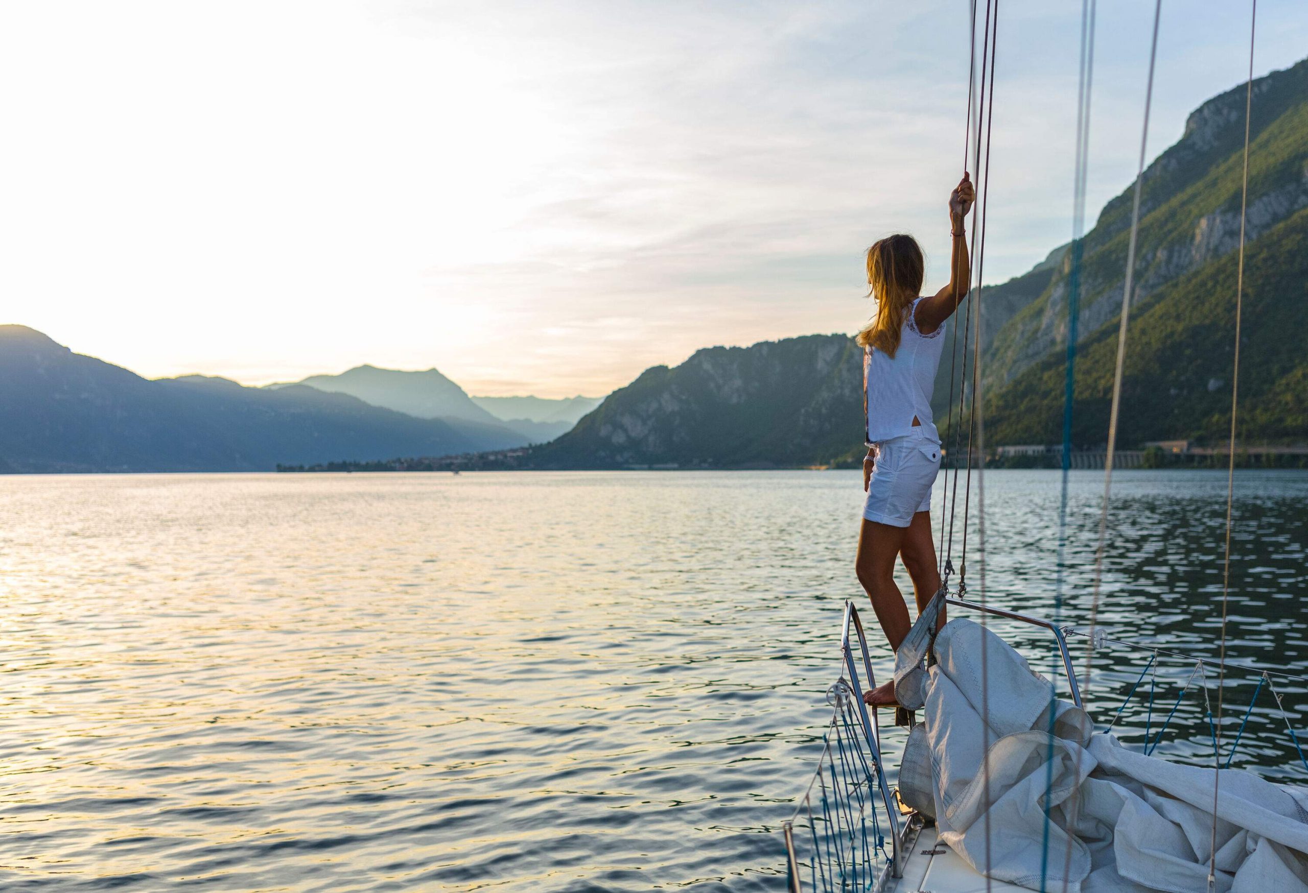 A person in a white shirt stands on the edge of a sailboat's front deck grabbing the ropes while gazing at the lake.