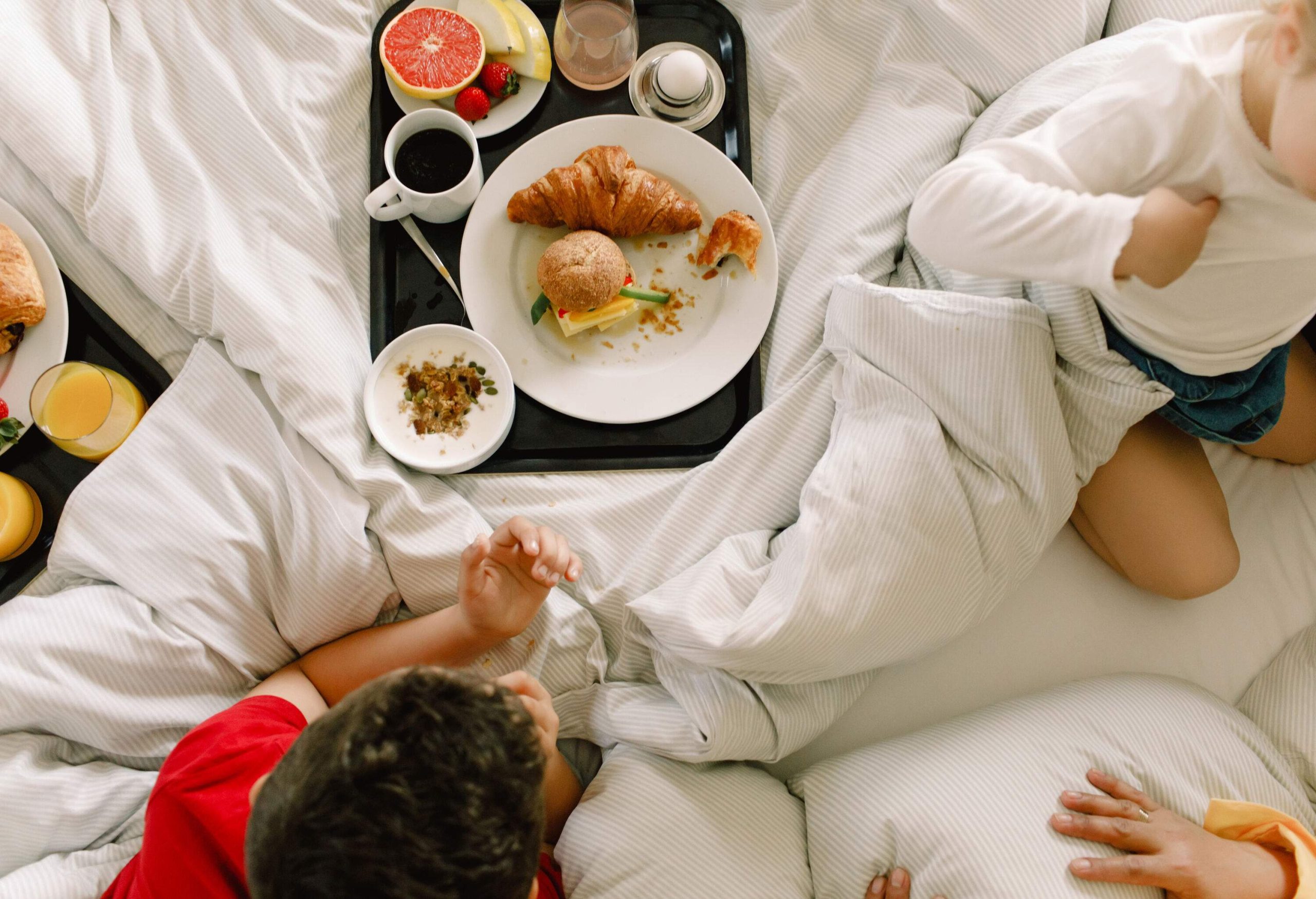 A group of kids on a bed with a tray full of burgers, pastries, cereal, coffee, and fresh fruits.