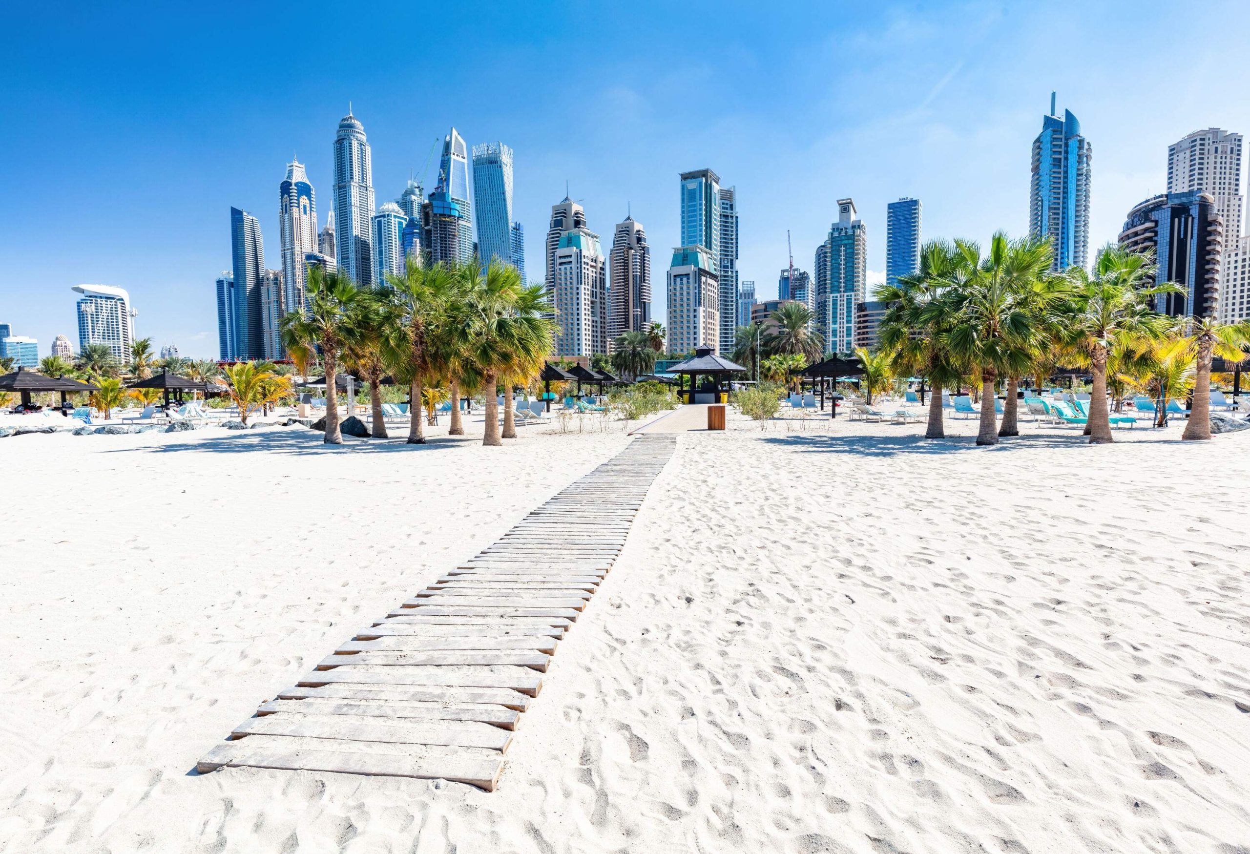 A wooden boardwalk runs through a sandy beach with palm trees and a backdrop of skyscrapers.