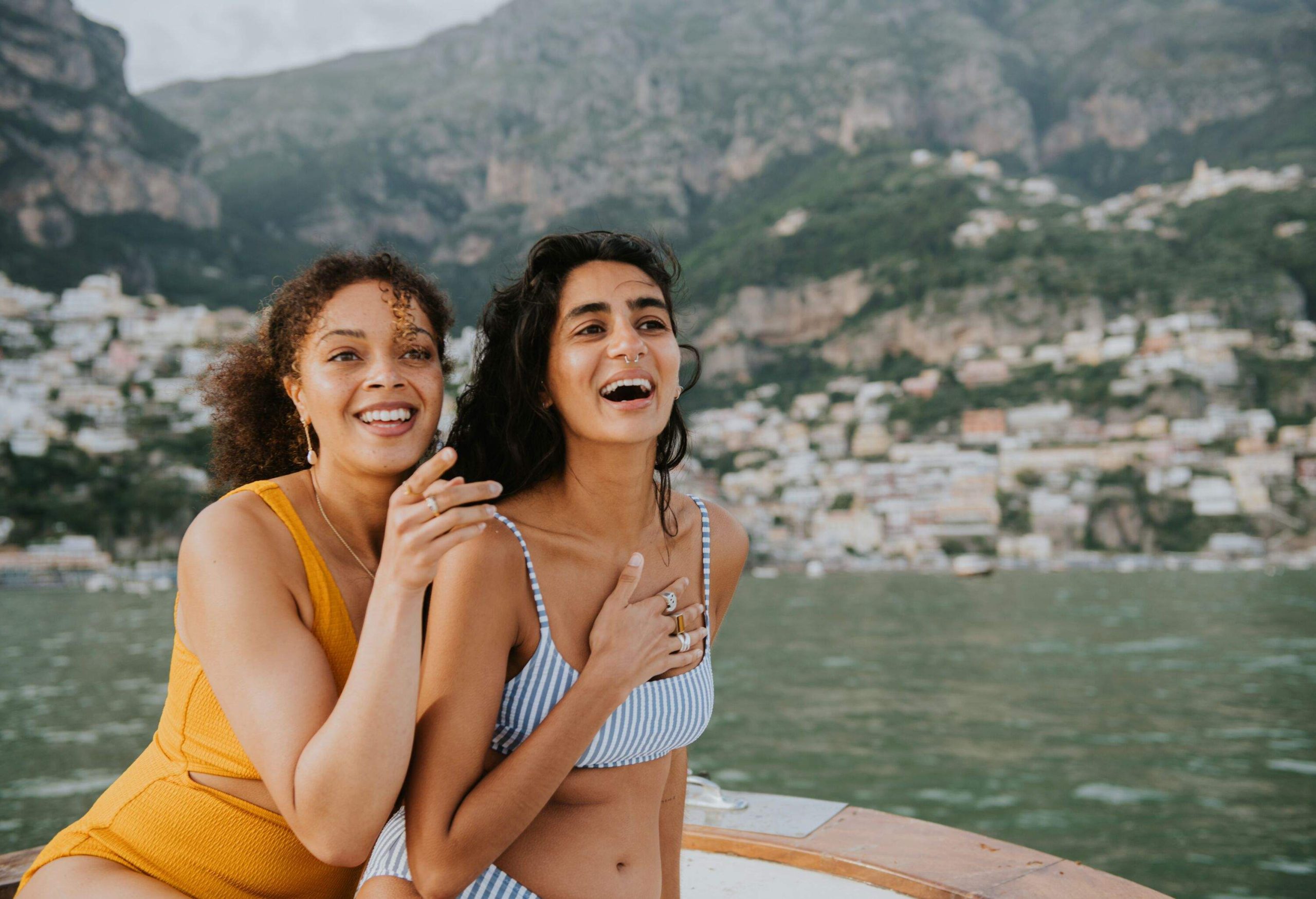 Two woman sit close together on the stern of a yacht. One points at something out of frame, and her friend looks surprised and delighted. View of Positano, Italy, visible in the background.