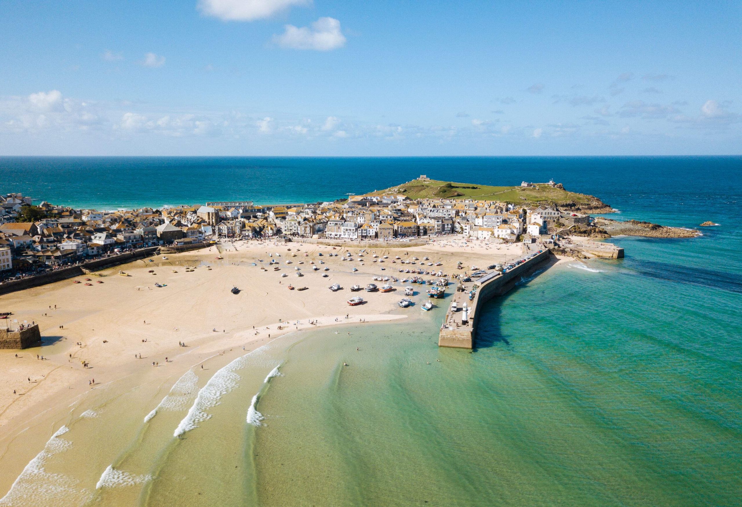 A sand beach with people strolling and relaxing near a breakwater and a settlement surrounded by the wide ocean.