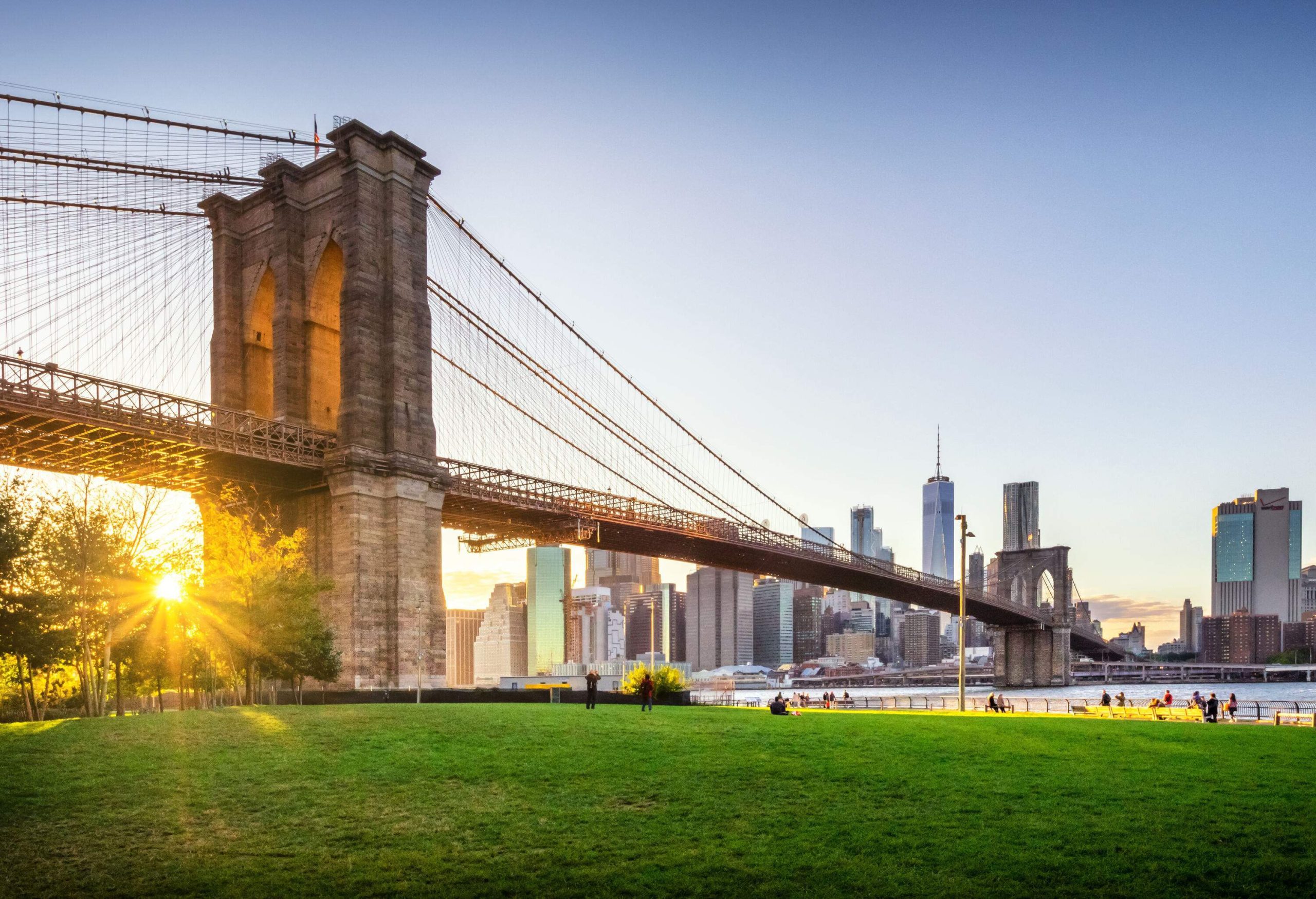 The long Brooklyn bridge spans the river between a grassy lawn and tall buildings.