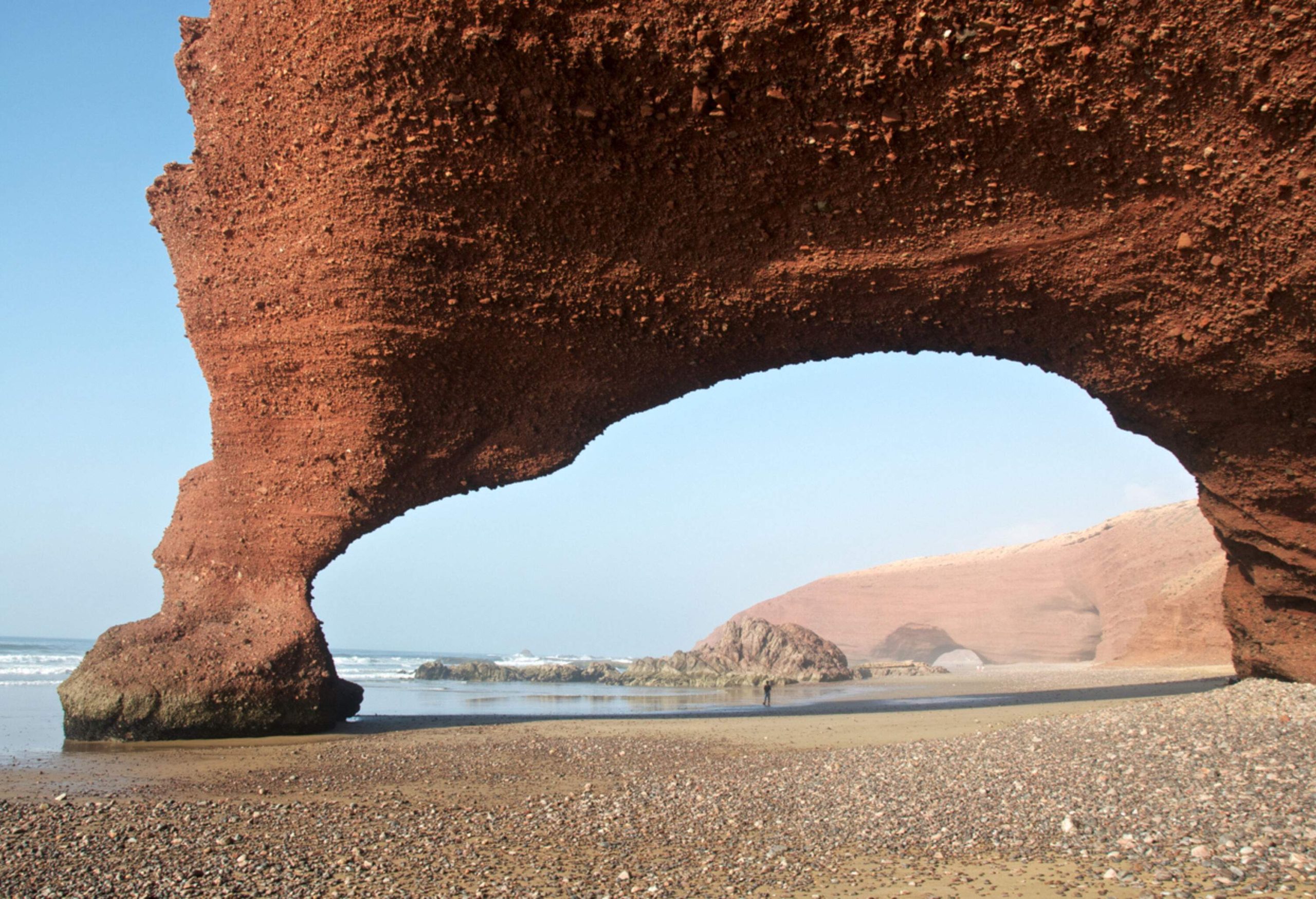 A person stands on a pebbled beach under an arched rock formation.