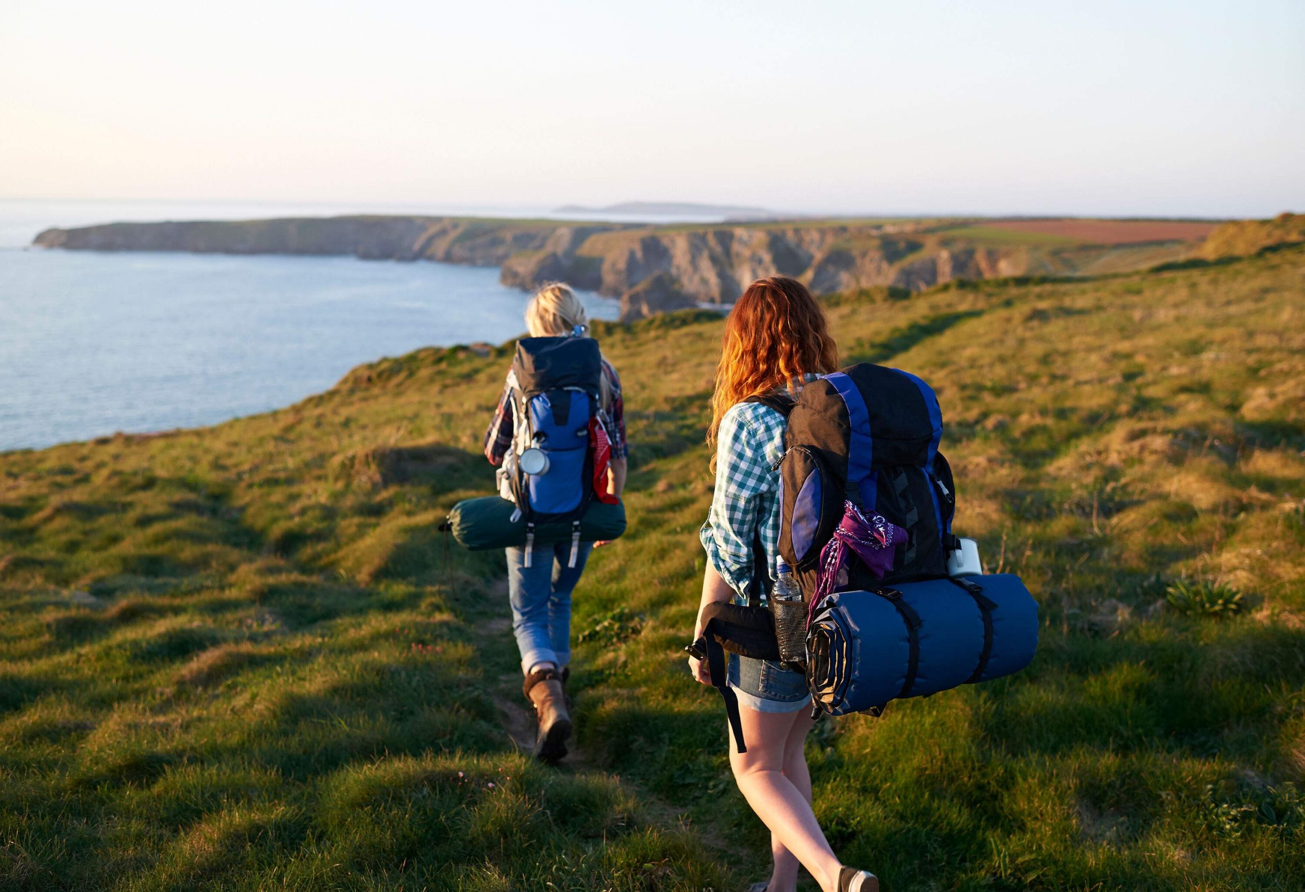 Two hikers carrying camping gear walk down a grassy dirt path with views of the coast in the distance.