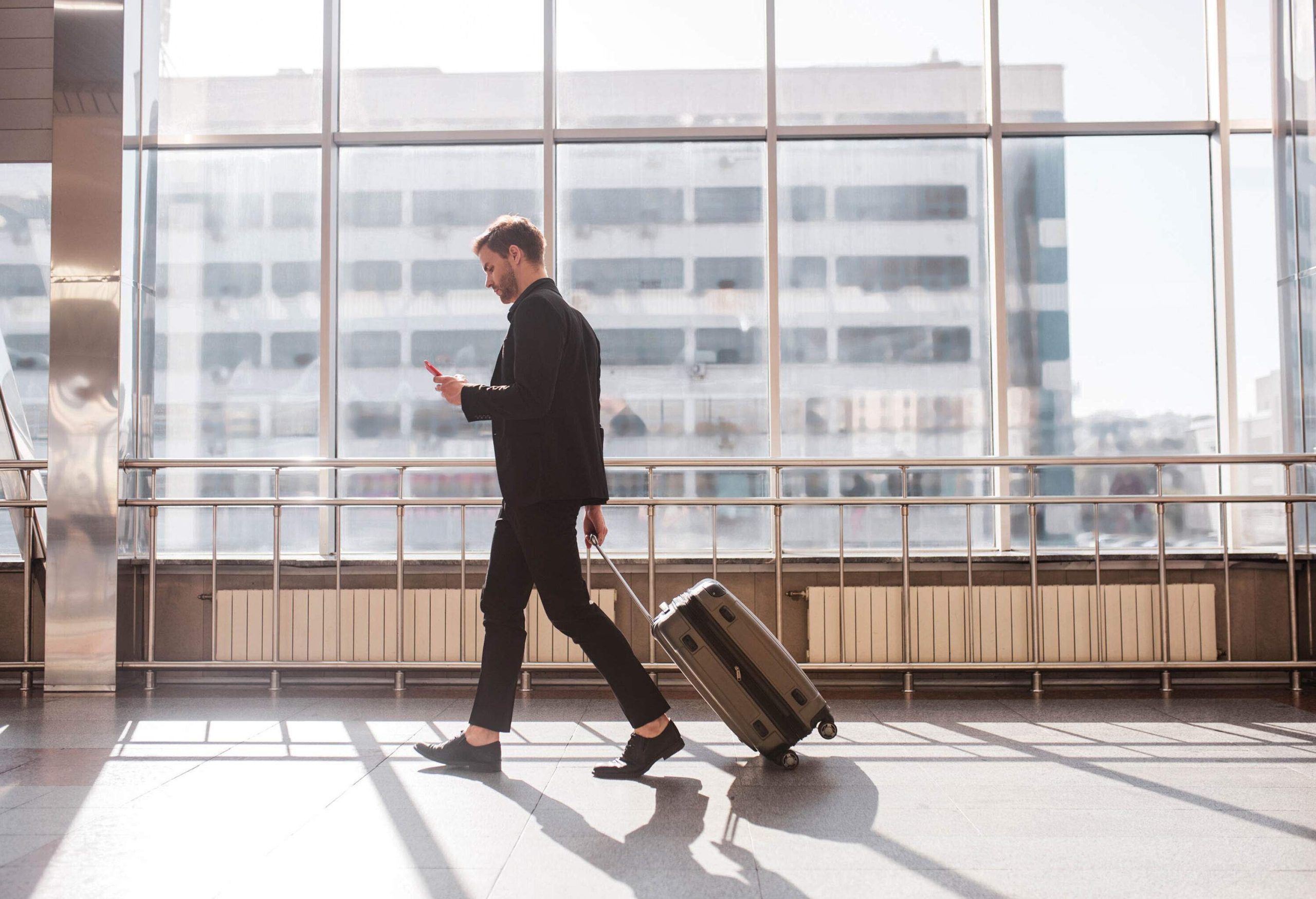 A male passenger uses his smartphone while walking with his luggage in the airport.