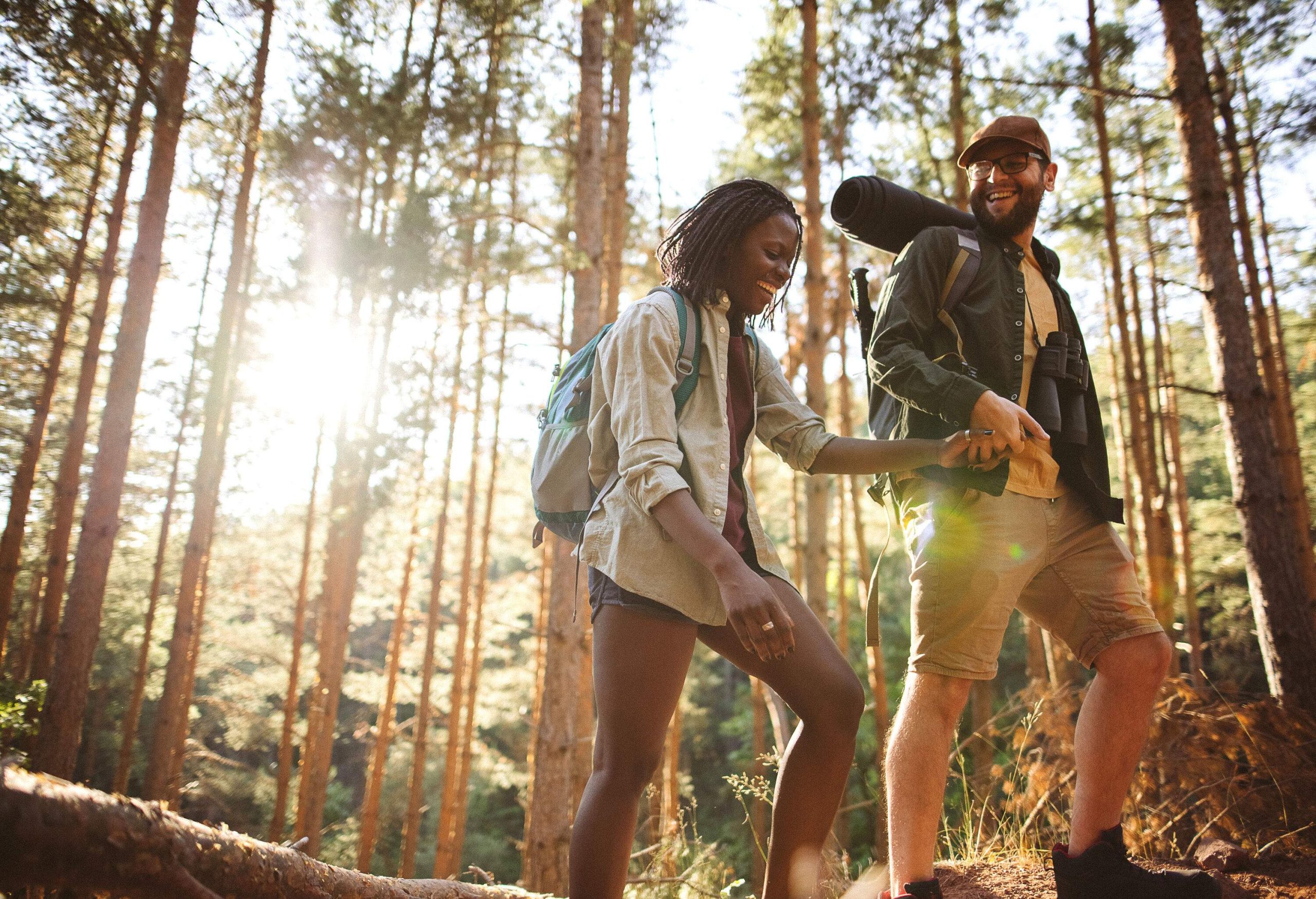 Happy couple strolls through the woodland holding hands while carrying camping bags.