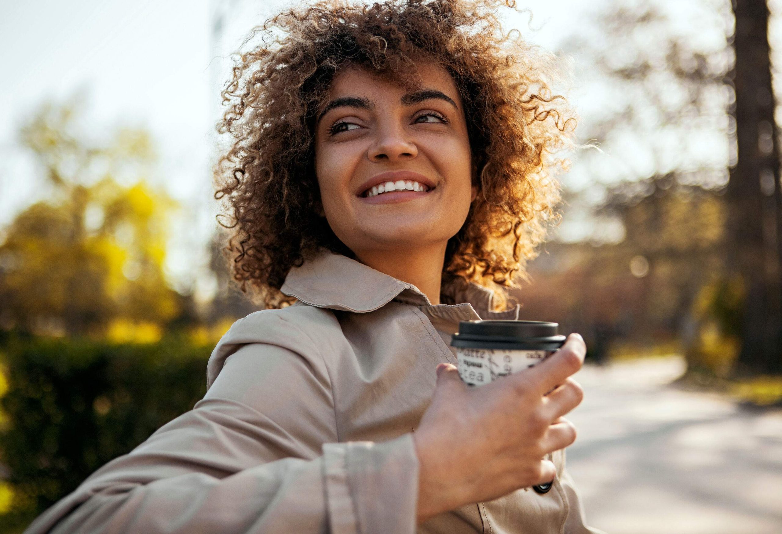 A curly lady holding a cup of coffee while walking in the park.