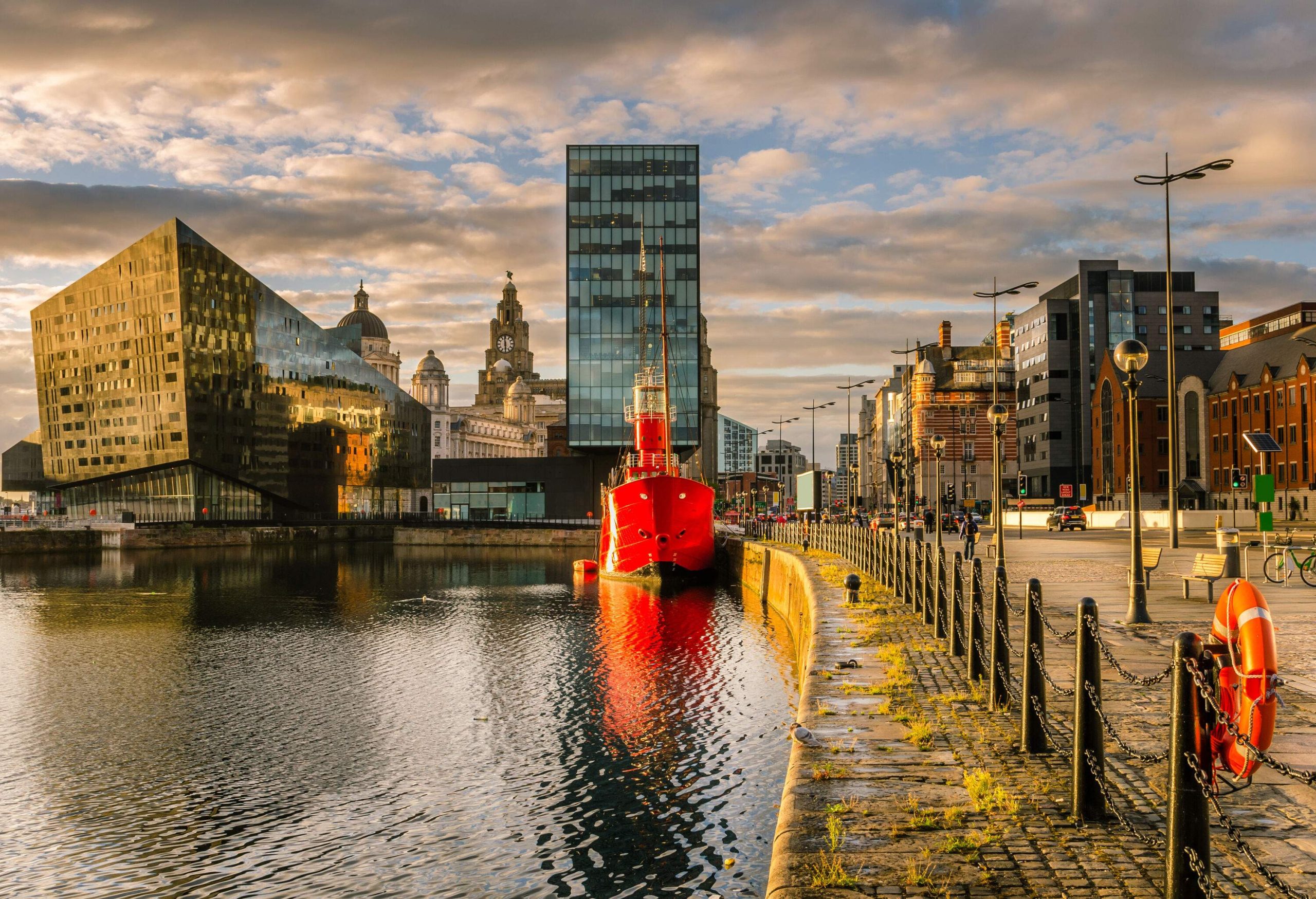 A red vessel docked in a harbour with both contemporary and historic houses lining the coast.