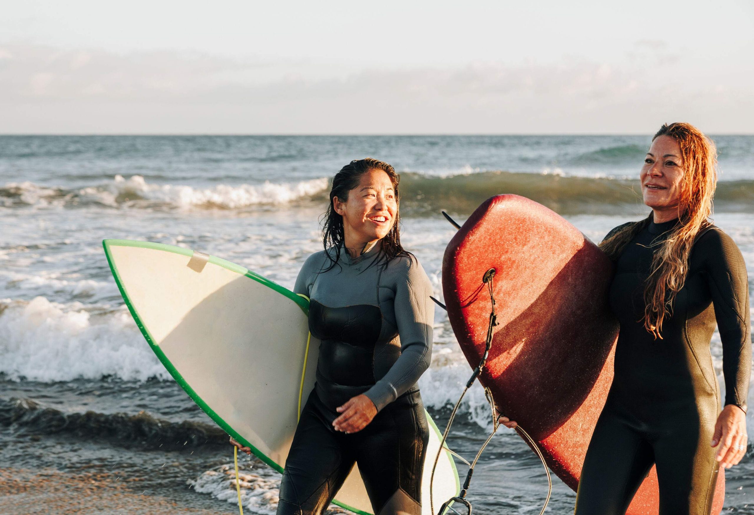 Two happy women in wetsuits holding surfboards on a wavy beach.