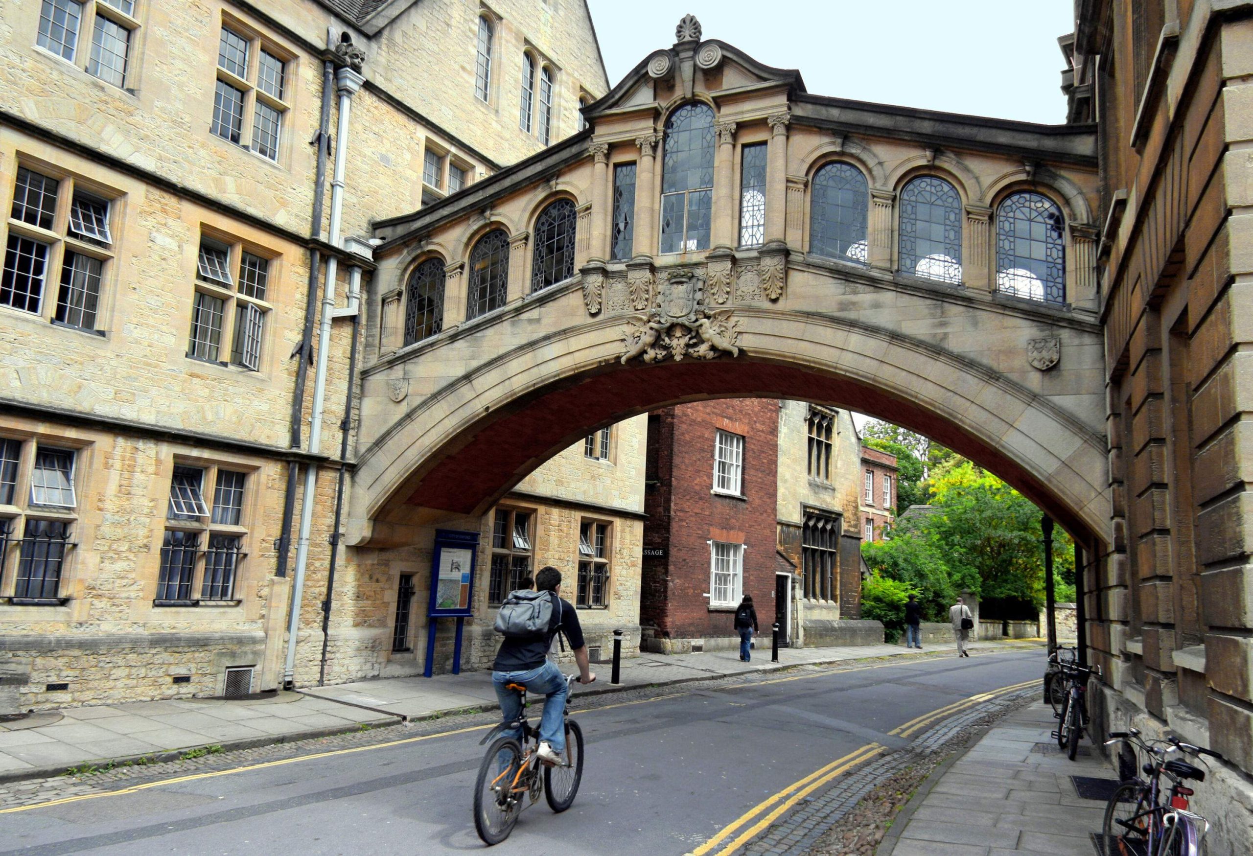 A cyclist on a single-lane street passes beneath a pedestrian bridge that spans a gap between two buildings.