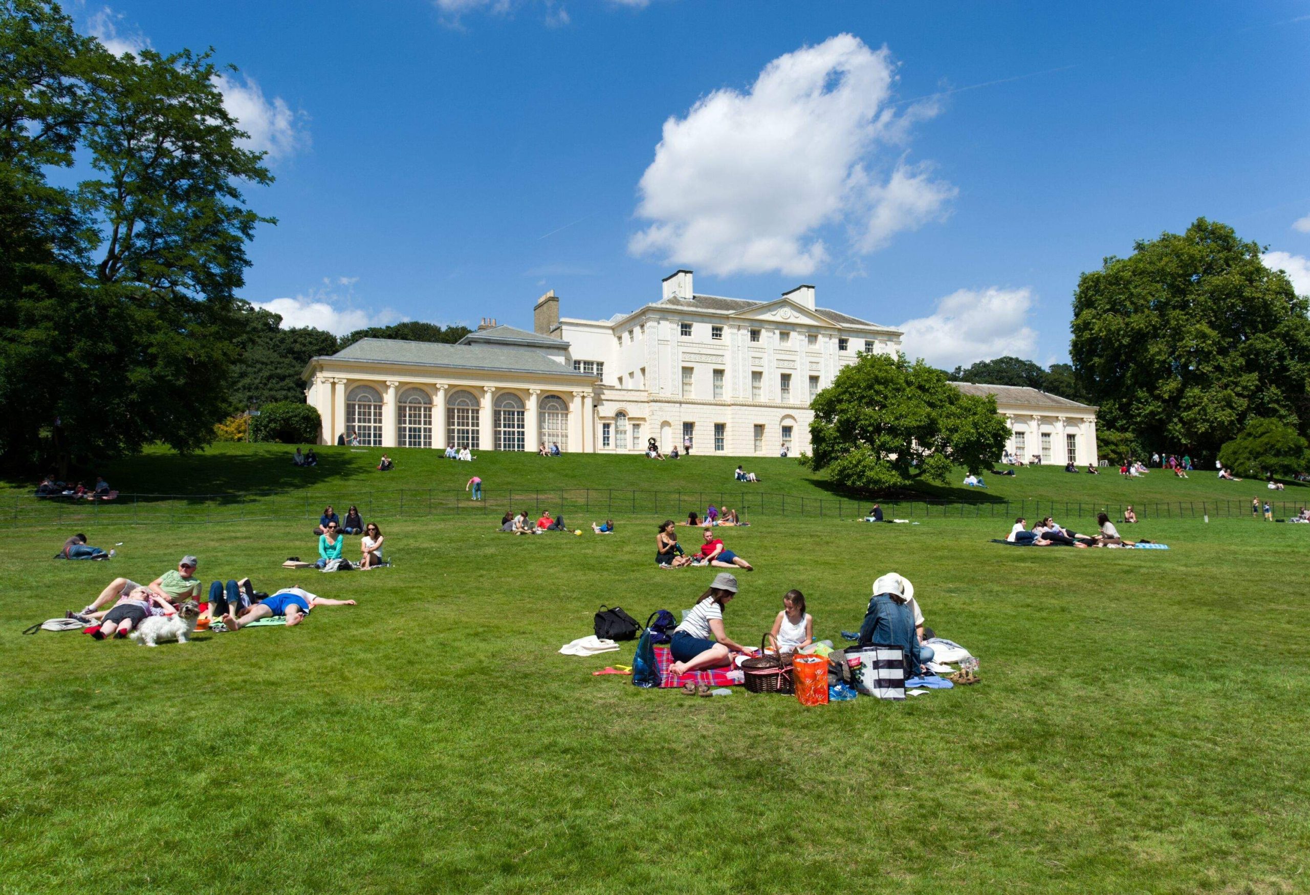 Individuals relish the sun in a verdant park in front of a classic white country house.