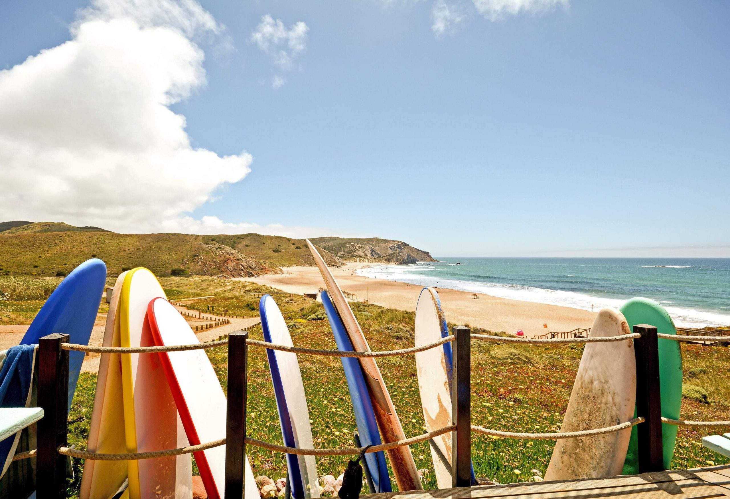 Colourful surfboards leaning against the rope fence of a deck overlooking the beach. 