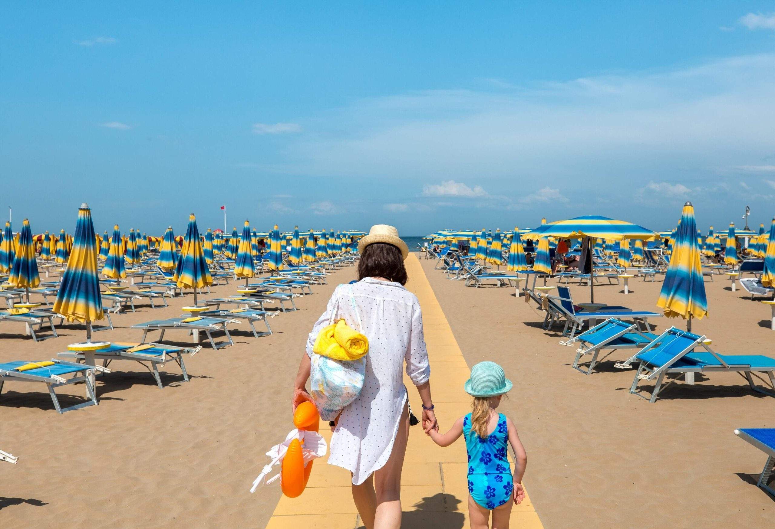 A woman holding a little girl's hand as they walk along the path across the beach strewn with umbrellas and loungers.