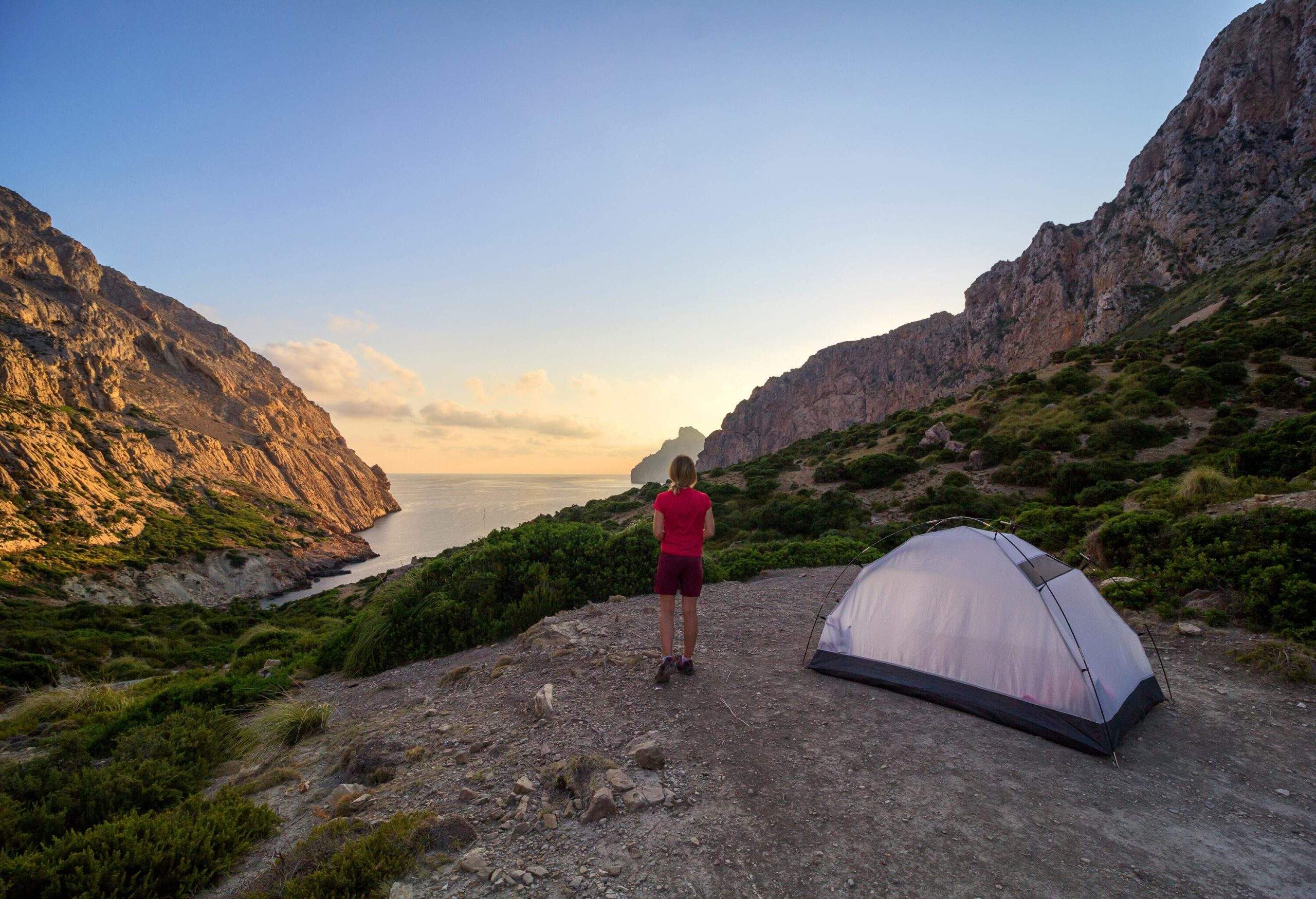 A lady watches the sunrise behind the rough mountains as she stands next to a white tent.
