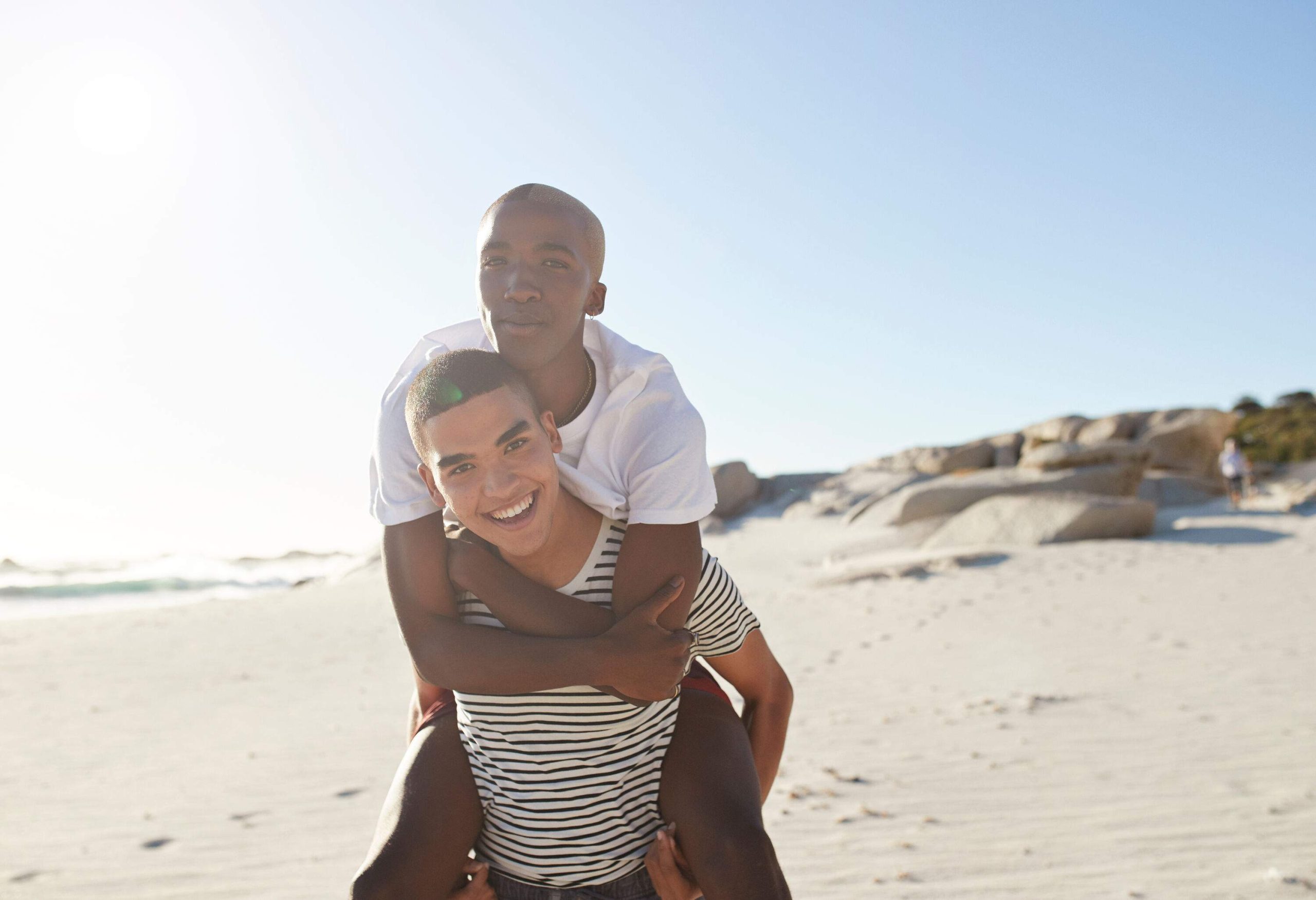 A man smiles as he gives a friend a piggyback ride on the beach.