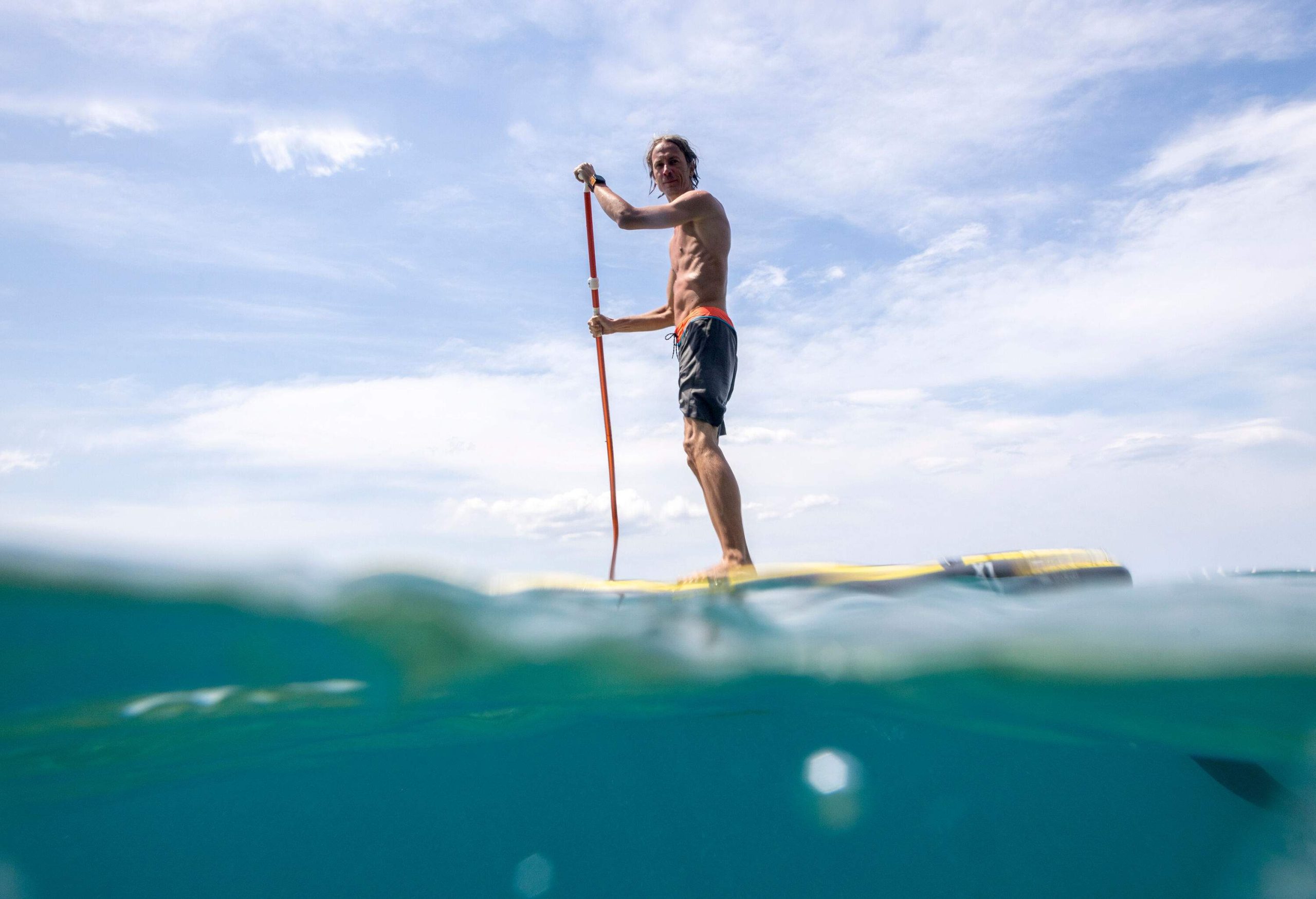 A man stands on a board propels through a paddle above the turquoise water.
