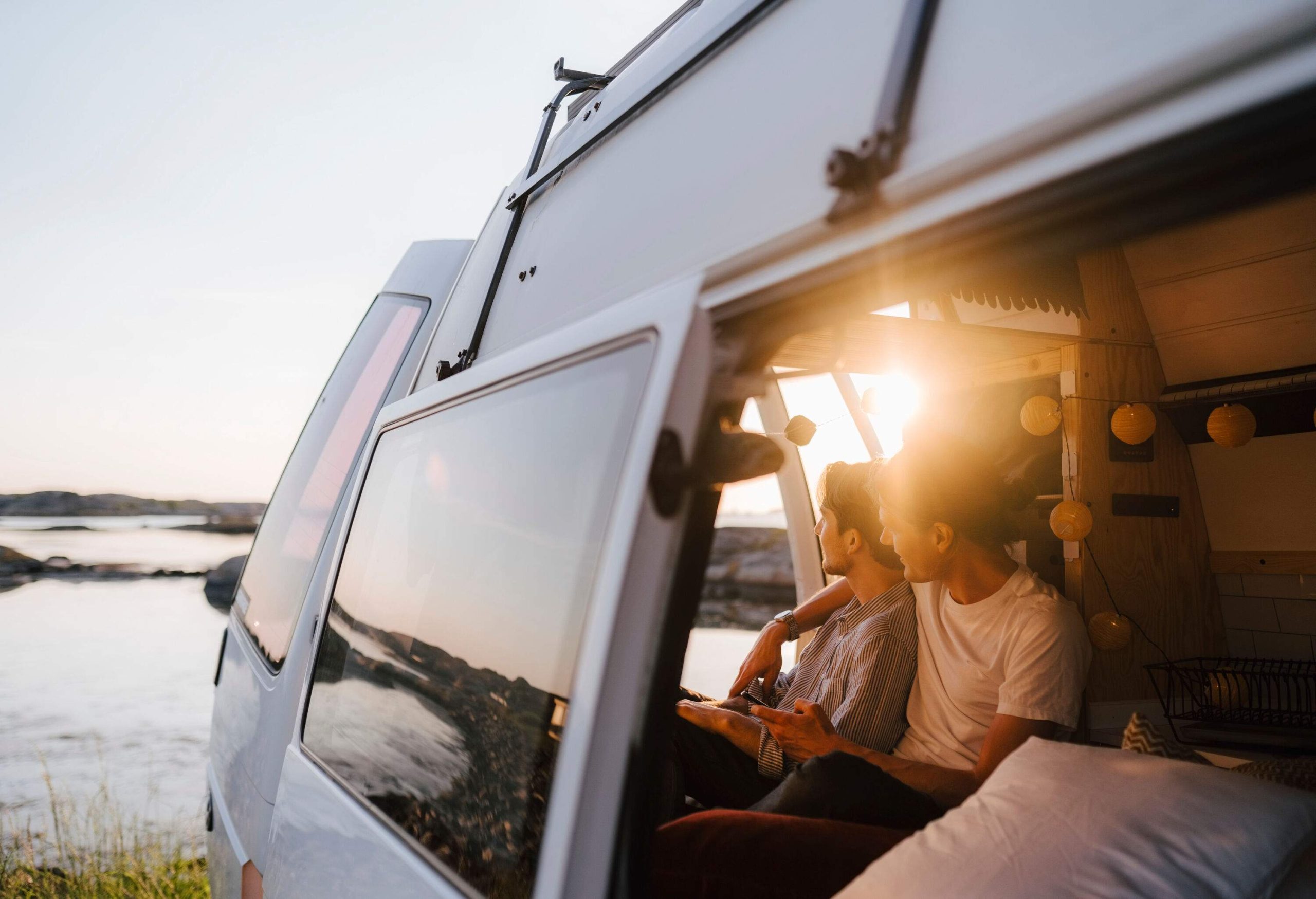Male couple sit in a camper van, one of them placing his arm around the other's shoulder as they look out the window at the surroundings.