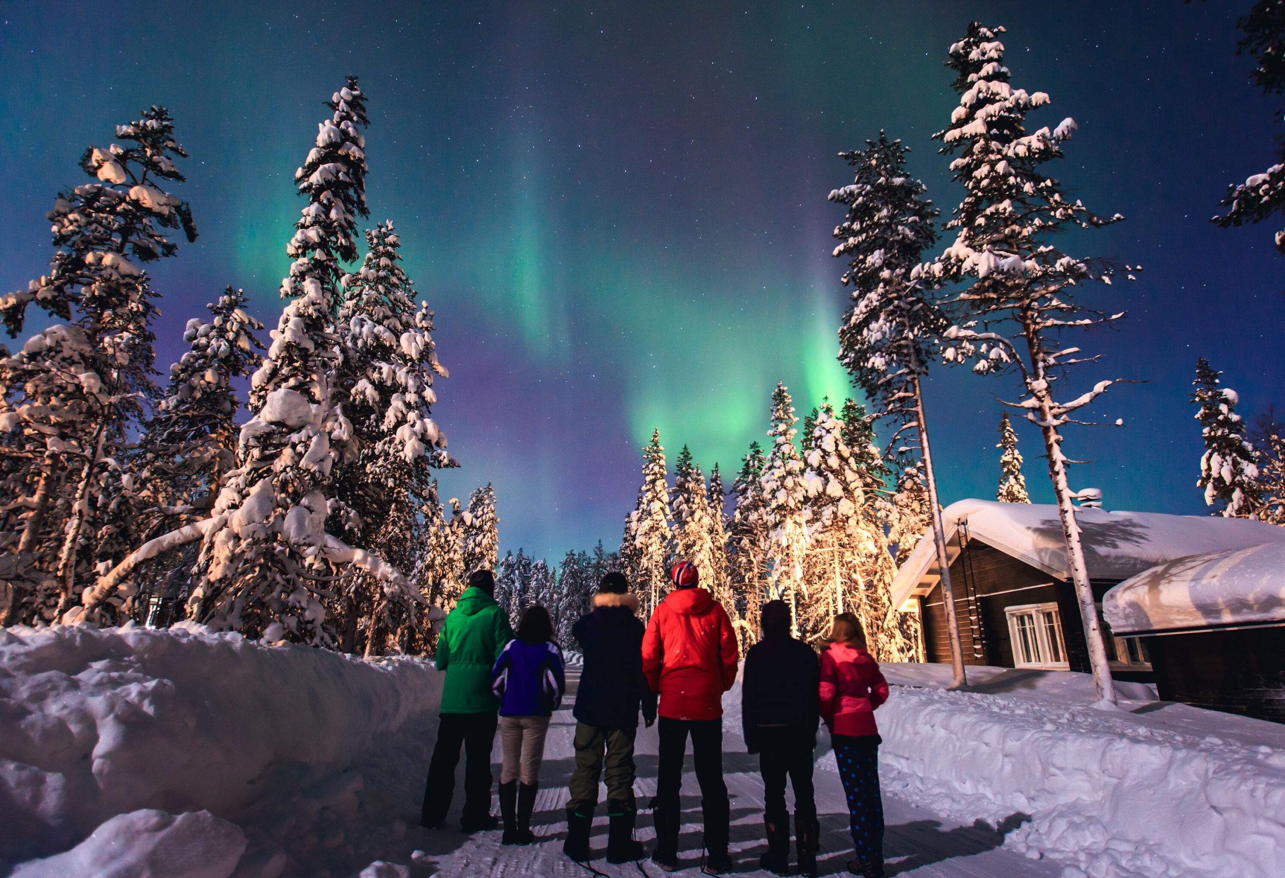 A group of friends lined up along a road to view the beautiful Northern Lights in a snow-covered village.