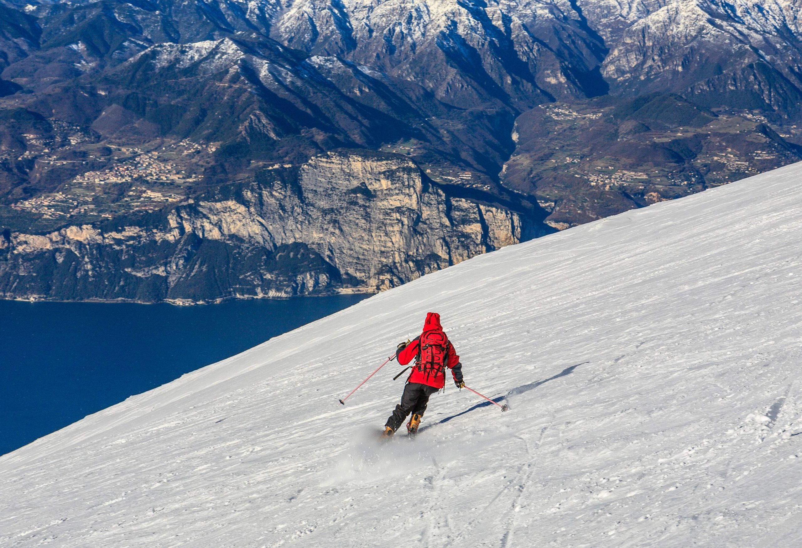 A skier in a red jacket gliding down a hill with a view of the blue lake and rocky mountains.