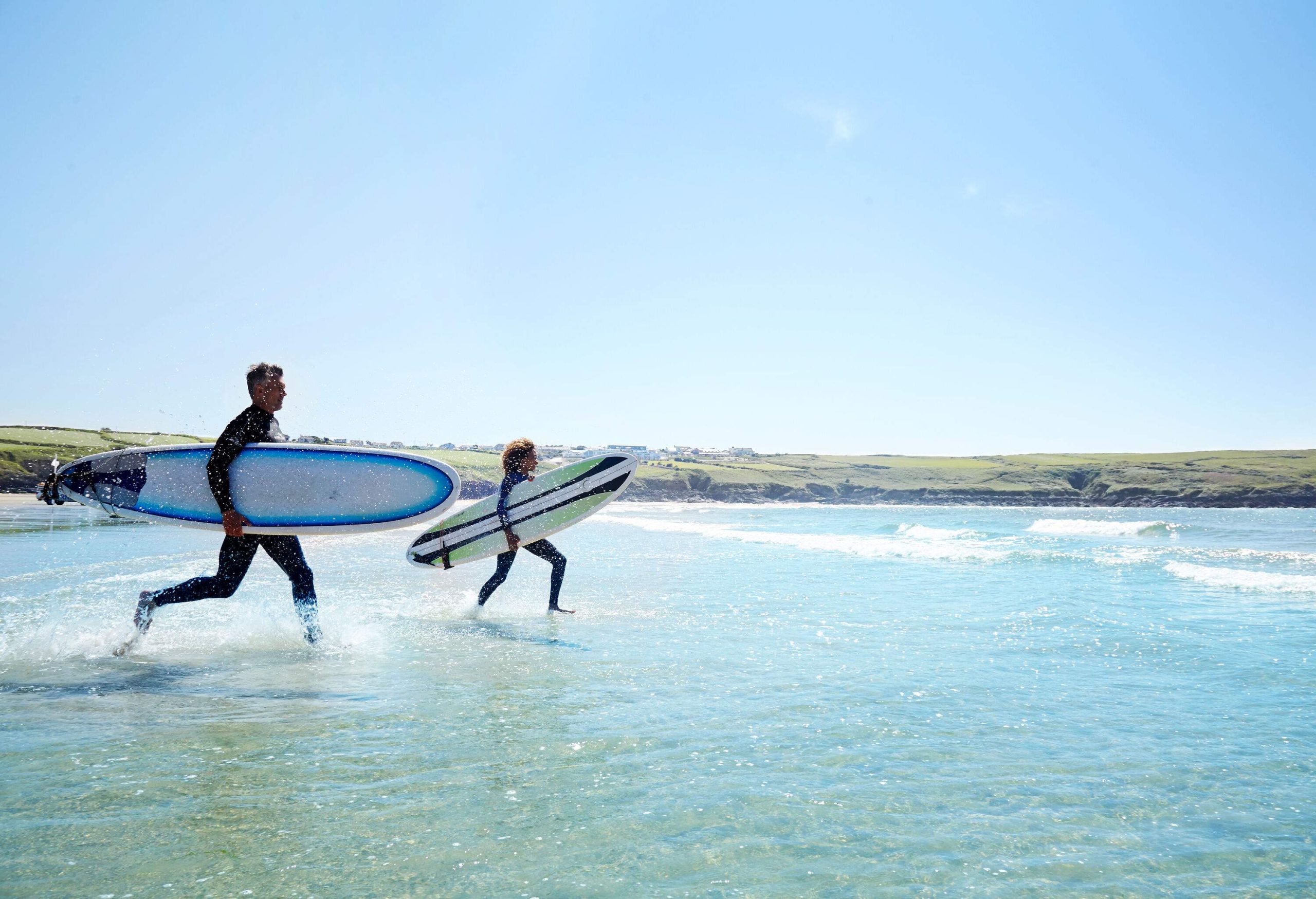Surfers with surfboards dash into the water against a backdrop of lush terrain.