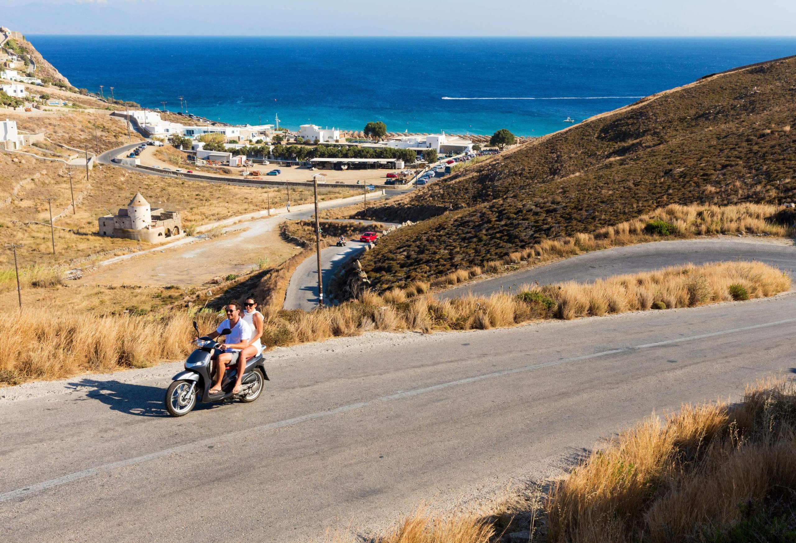 A couple riding on a scooter traveling on a roadway across a hill with the sea in the background.
