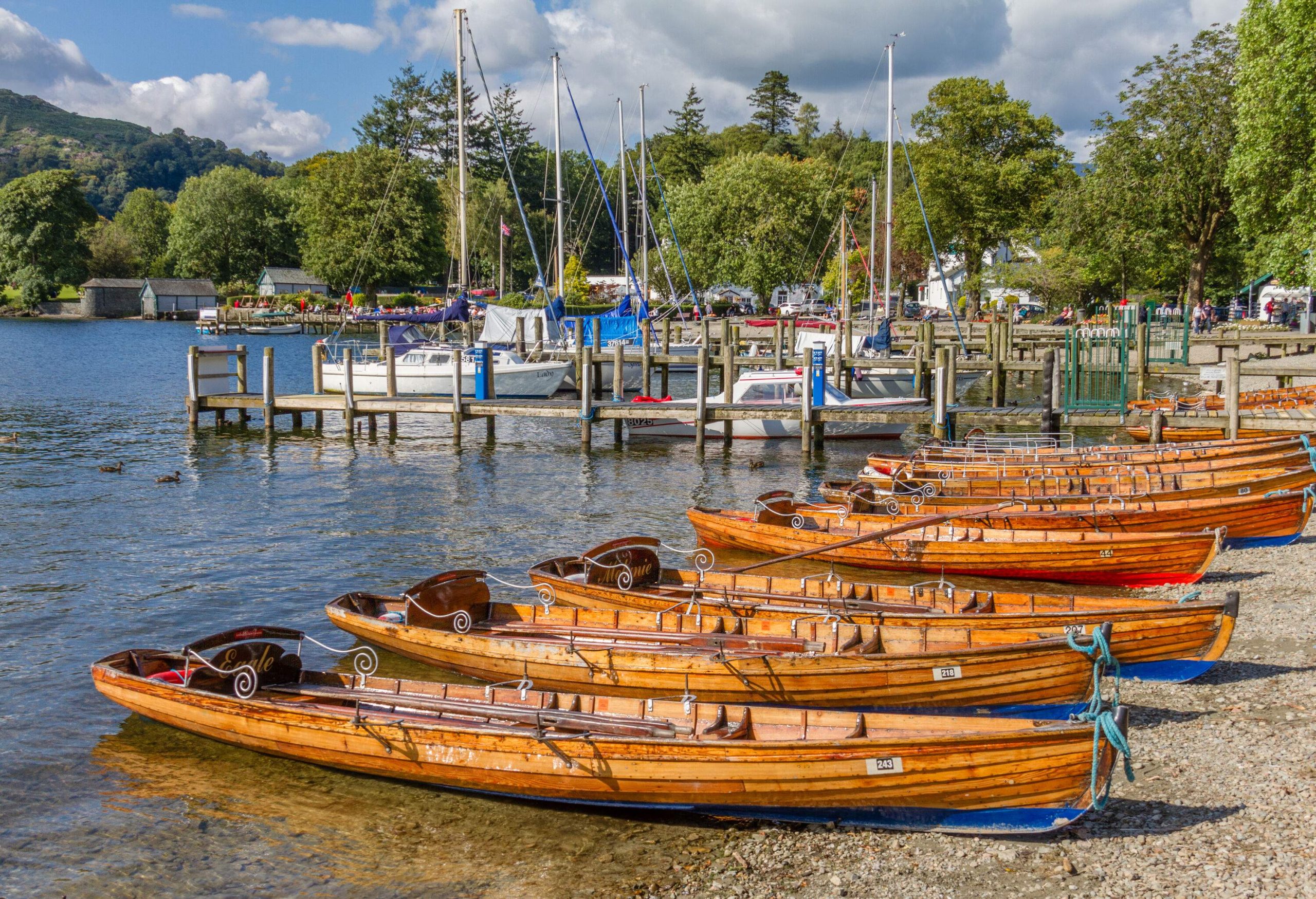 A line of orange rowing boats parked on a beach next to a pier with anchored sailboats.