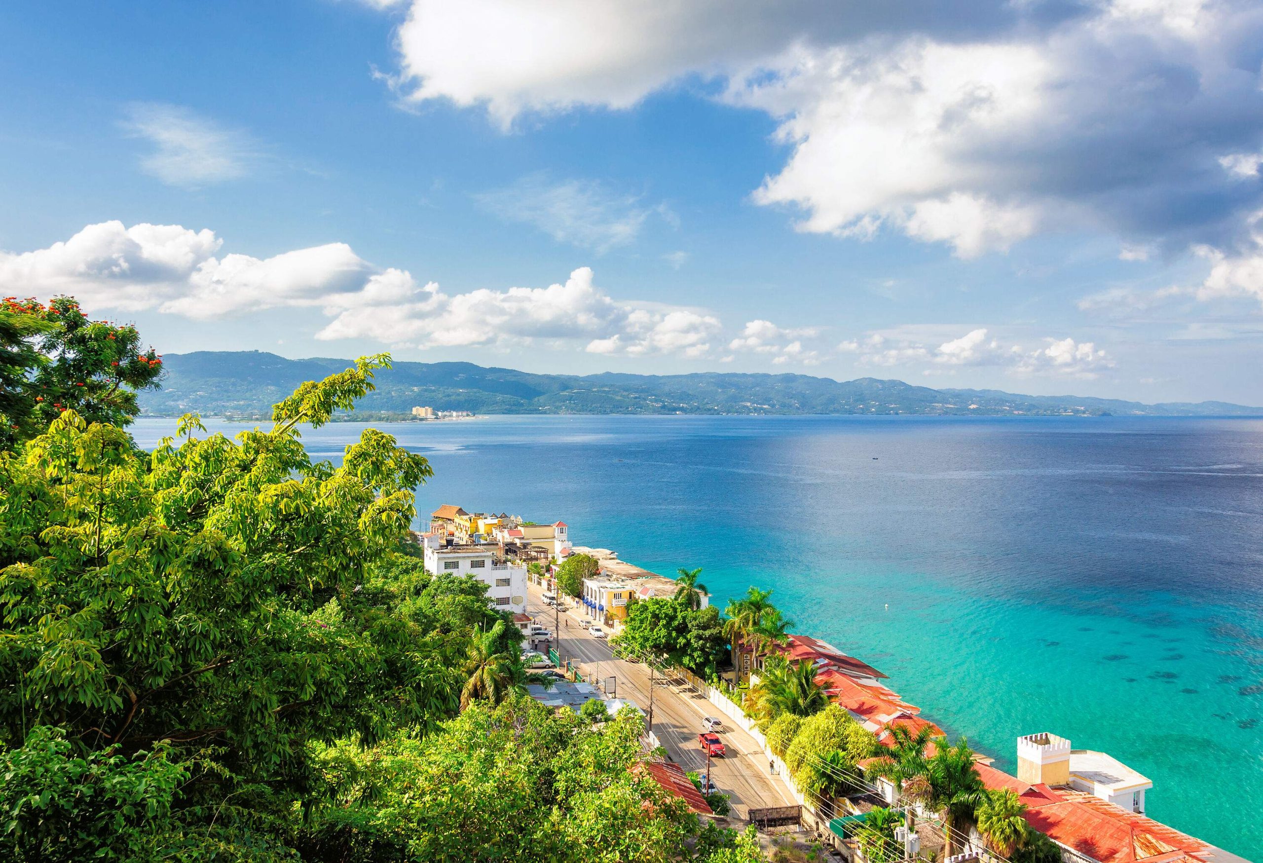 Aerial view of a lush coastal town overlooking the tranquil turquoise sea with the mountain range against the cloudy blue sky.