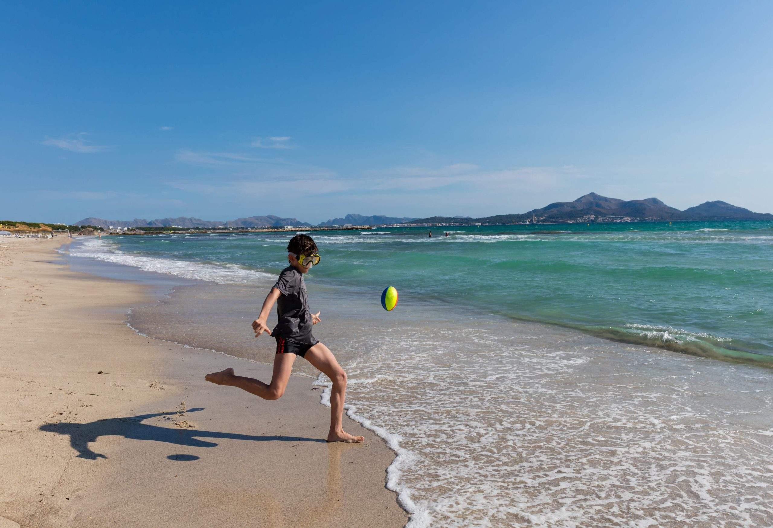 A little boy wearing goggles playing with a rugby ball on the beach.
