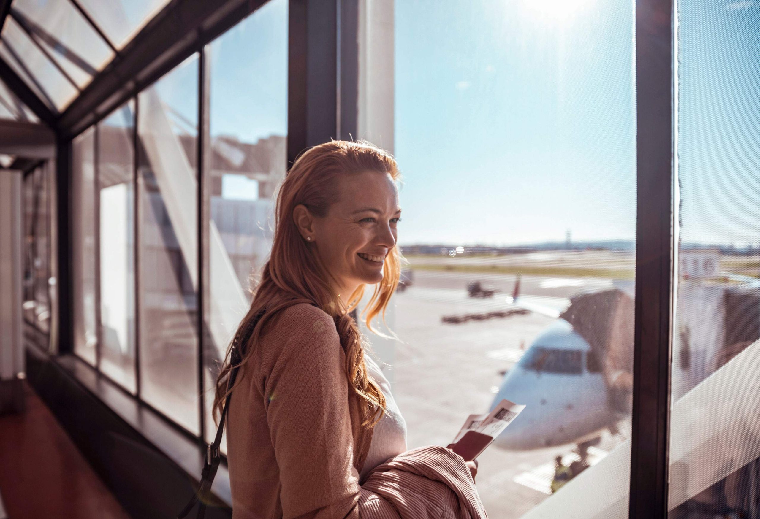 A woman smiles as she stands beside the big glass windows of an airport terminal while holding her ticket.