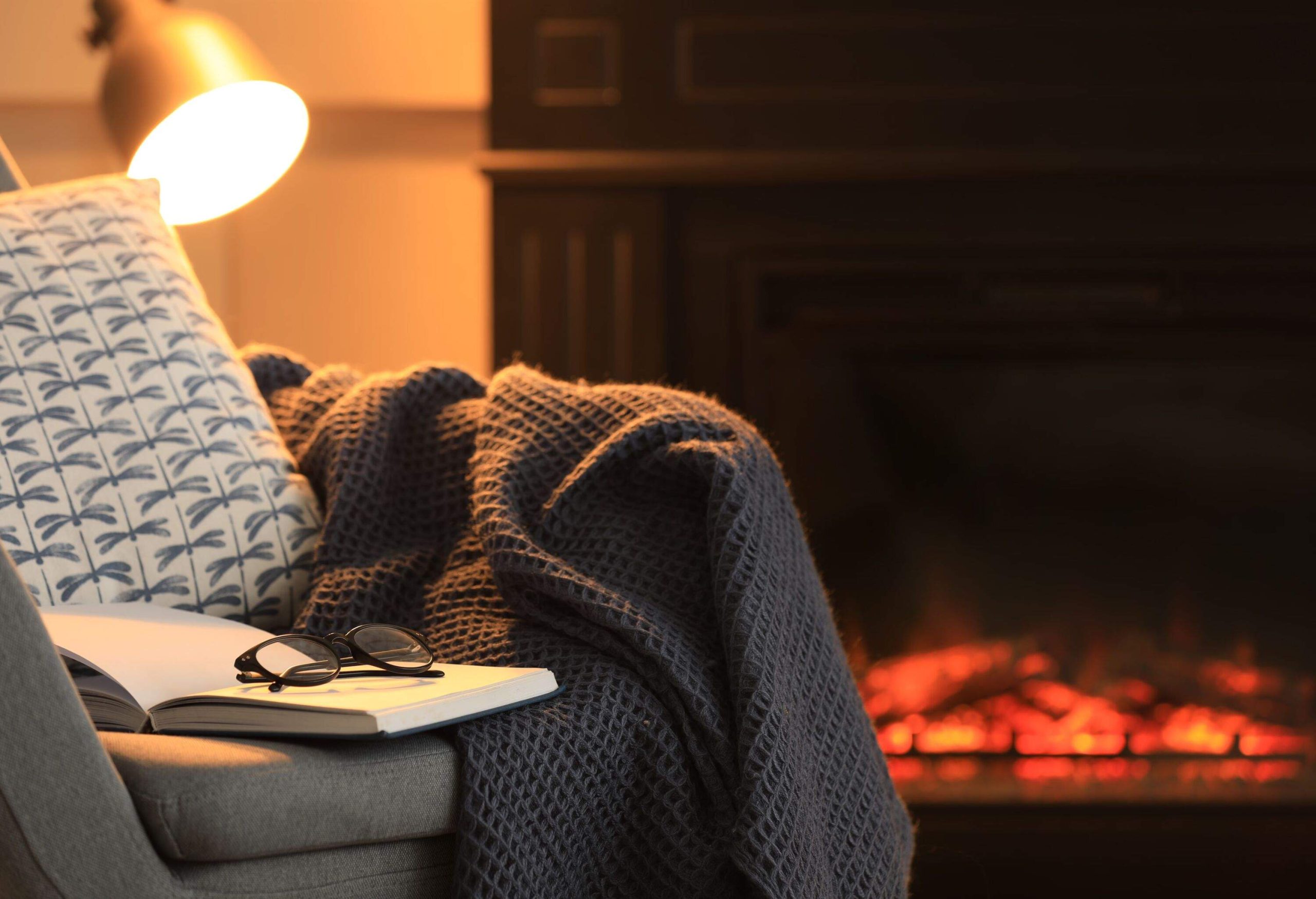 Open book, glasses and plaid on armchair near fireplace at home, space for text. Cozy atmosphere