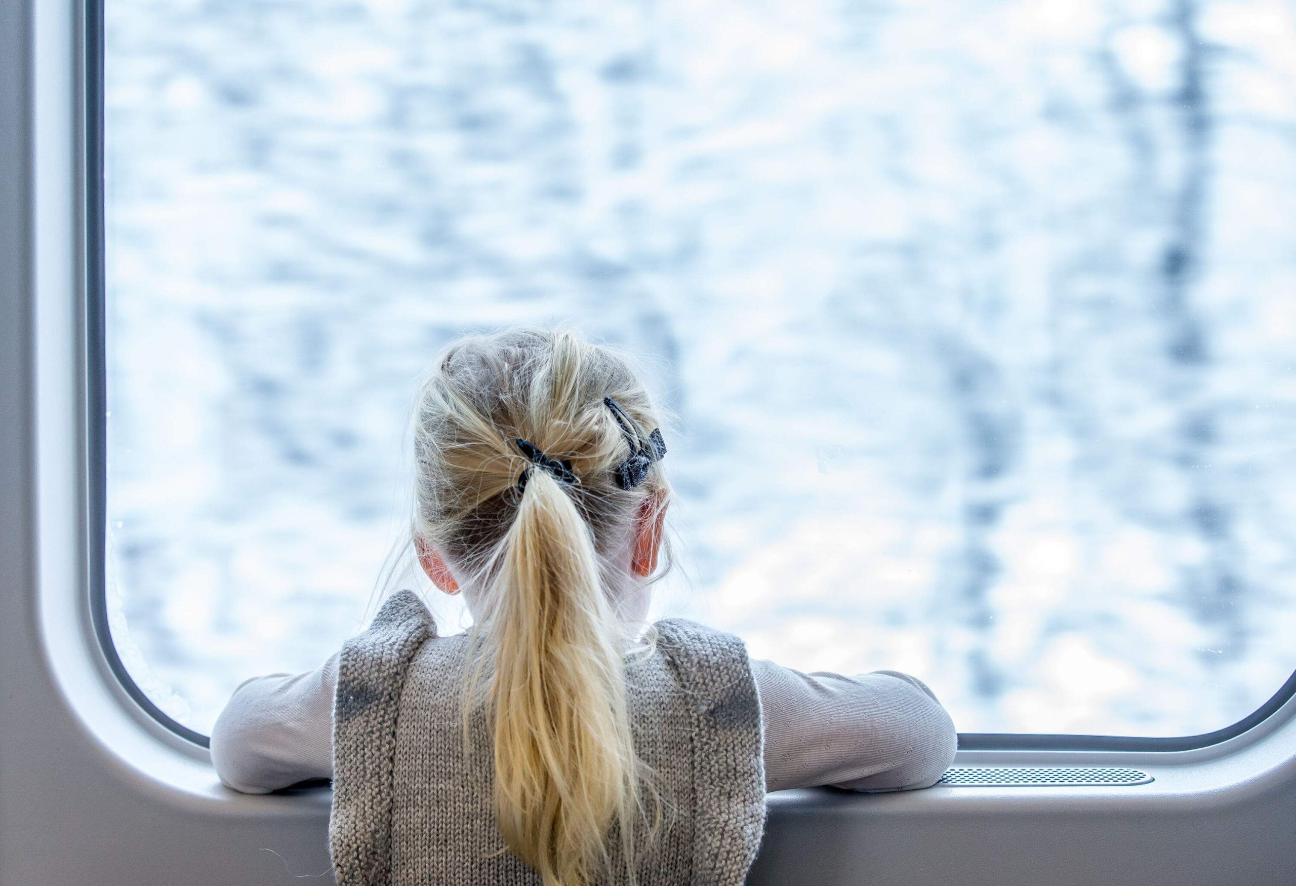 A blonde girl looks out a train's window.